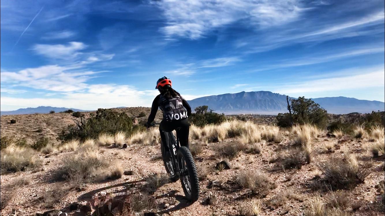 A person riding a mountain bike on a dirt trail surrounded by desert vegetation, with mountains and a blue sky in the background. The cyclist is wearing a helmet and a black shirt with "VOM" printed on it. Mariposa Fat Bike Trails mountain bike trail.