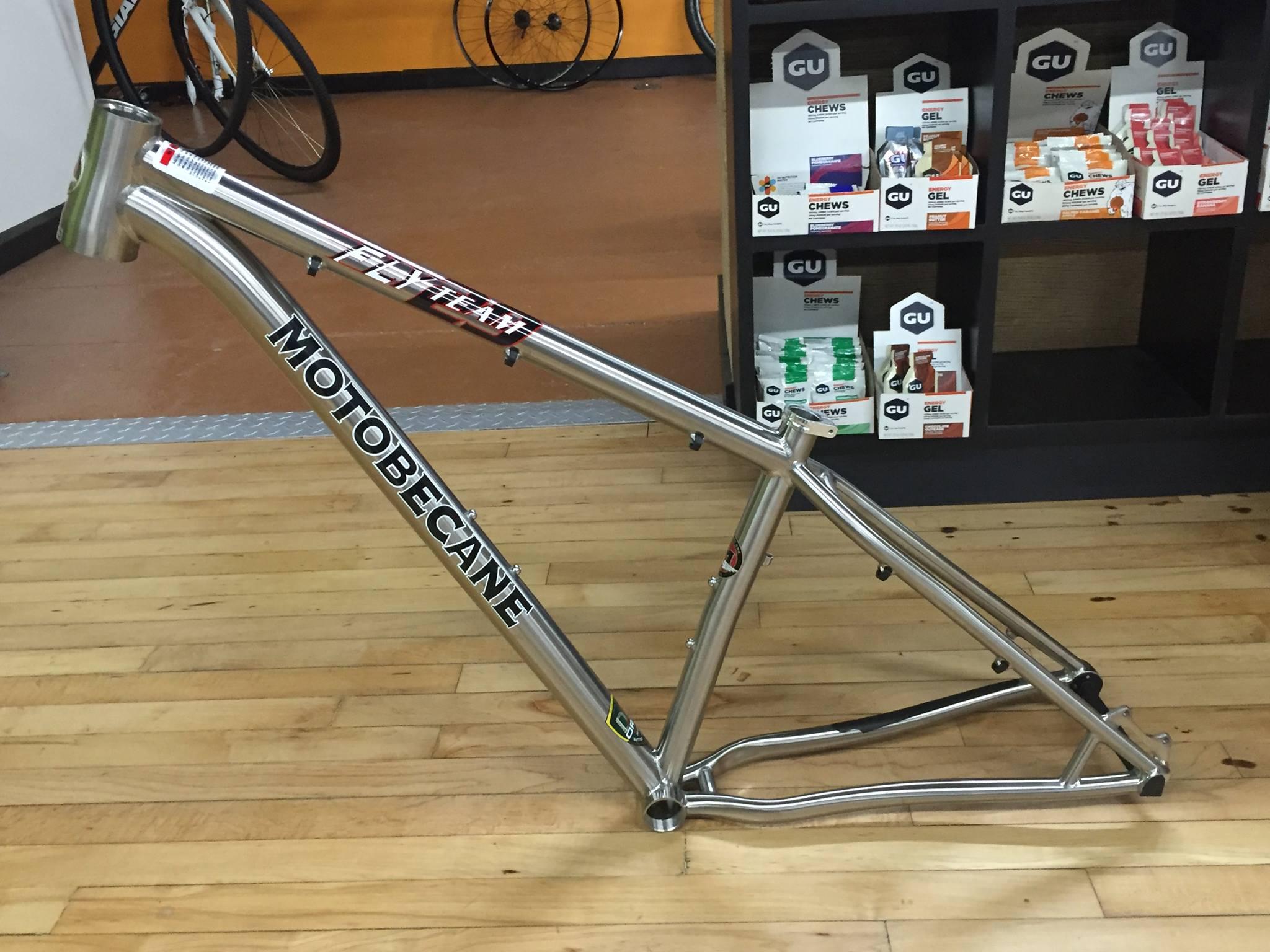 Motobecane Fly Team Ti 29er: A silver Motobecane bicycle frame displayed in a retail setting, with a wooden floor and shelving in the background containing cycling gel products. The frame features branding details with the word "MOTOBECANE" prominently displayed.
