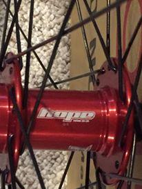 Motobecane Fly Team Ti 29er: Close-up image of a red bicycle hub with the brand logo "Hope" visible. The hub is part of a wheel assembly, showcasing the spokes and a textured background.