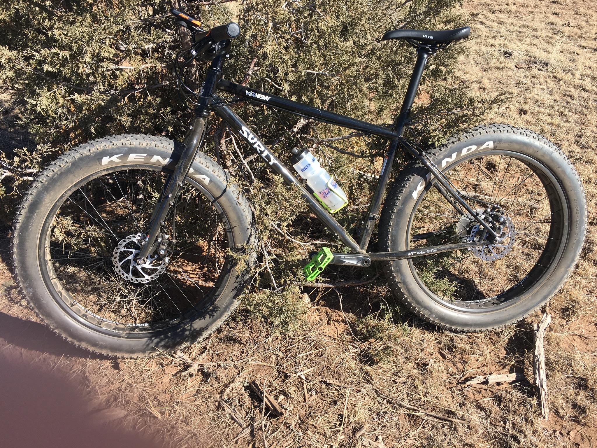 Surly Wednesday: A black fat bike leaning against a bush, featuring wide tires marked with "KENDA." The bike has disc brakes, a green pedal, and a water bottle holder attached to its frame. The background shows dry grass and earth.