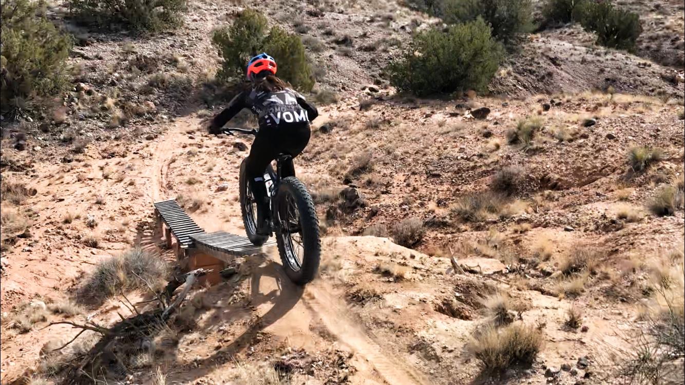 A mountain biker in a black outfit and bright orange helmet is navigating a narrow wooden plank bridge on a trail in a rocky, desert landscape. The bike features wide tires, and the surrounding area is dotted with shrubs and natural terrain. Mariposa Fat Bike Trails mountain bike trail.