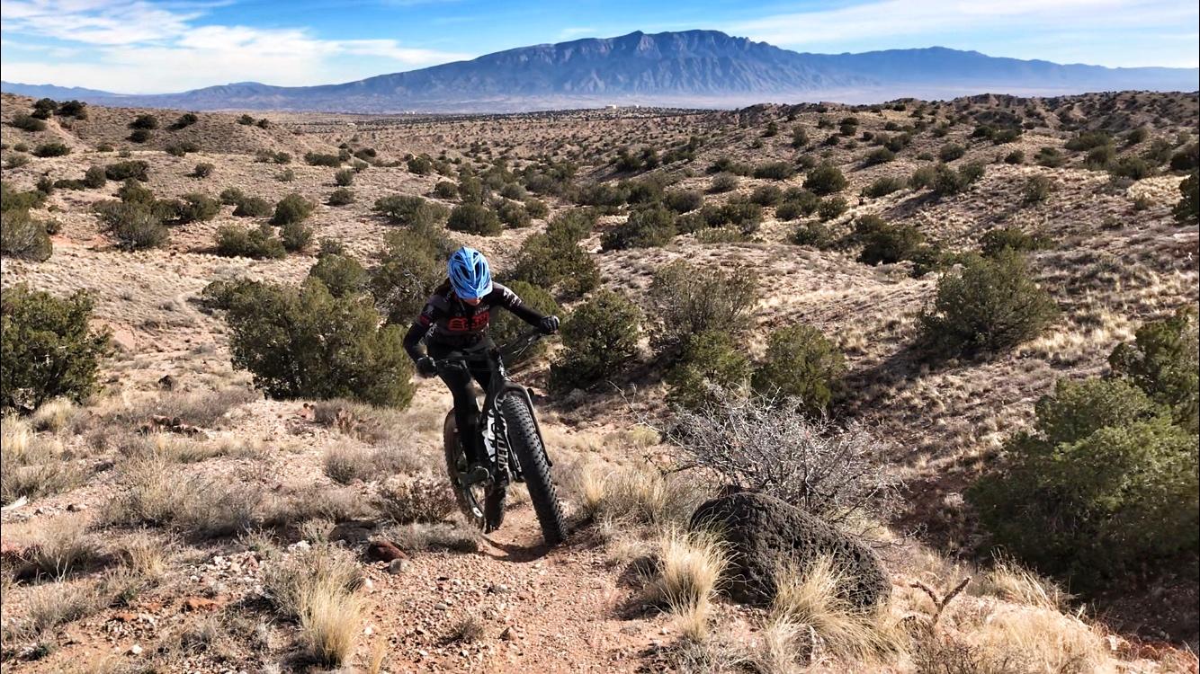 A cyclist riding a fat bike on a rocky trail in a desert landscape with shrubs and hills, while mountains are visible in the background under a clear blue sky. Mariposa Fat Bike Trails mountain bike trail.