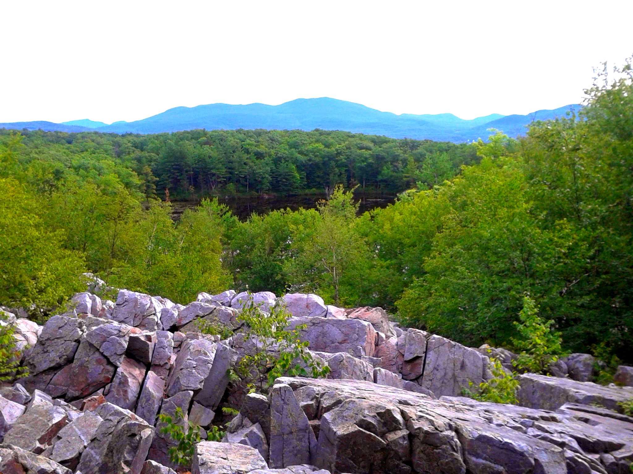 A scenic view of rocky terrain in the foreground with lush greenery and a forest in the midground, leading up to rolling mountains in the background under a bright, overcast sky. Pine Hill Park mountain bike trail.