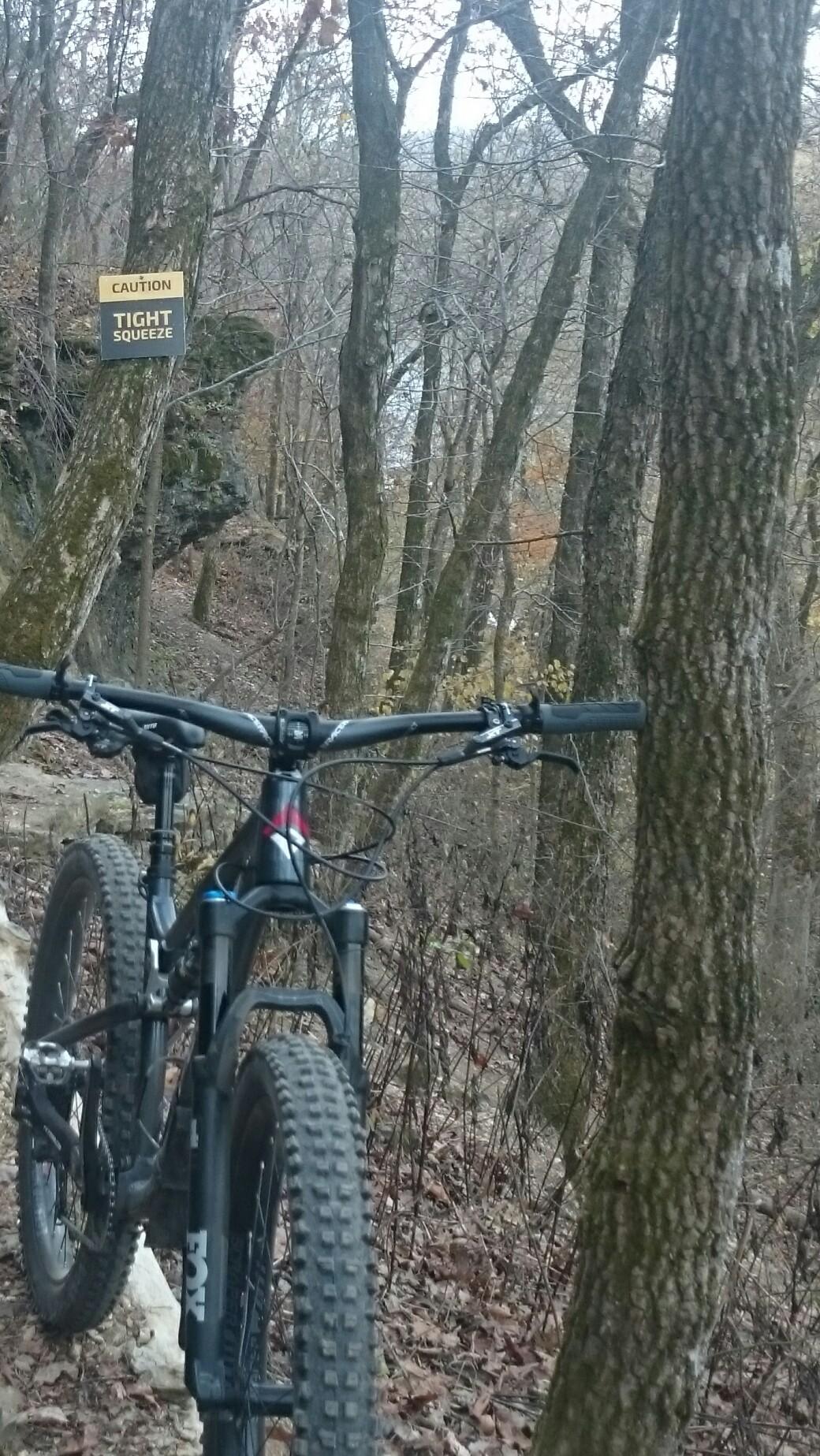 Fezzari Cascade Peak: A mountain bike positioned on a narrow trail surrounded by trees, with a "Caution: Tight Squeeze" sign visible in the background. The scene conveys a rugged outdoor setting in a forested area during autumn.