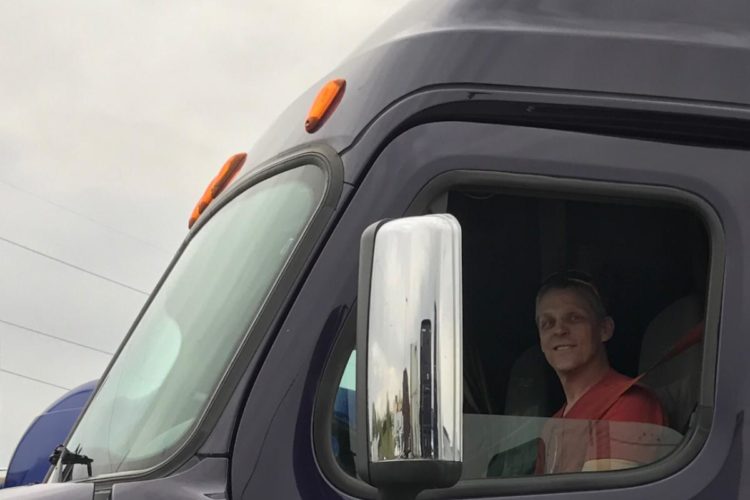 A smiling truck driver sitting in the cab of a large, dark-colored truck. The reflection in the side mirror shows part of the truck's exterior and surroundings. The sky is overcast, adding a gray backdrop to the scene.