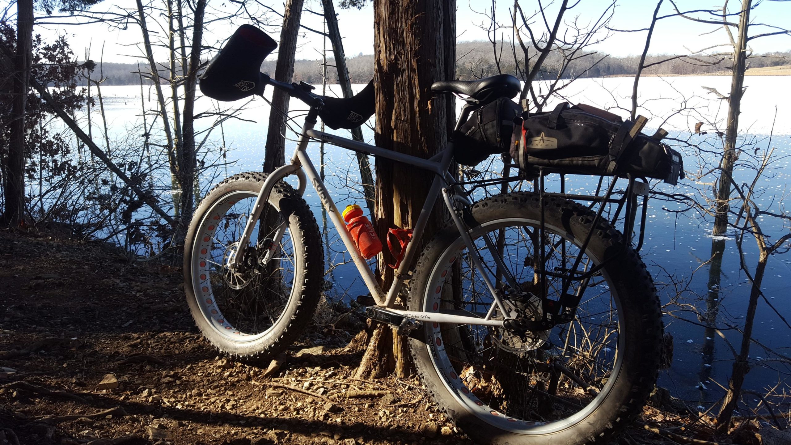 A fat tire bicycle leaning against a tree near a calm body of water, surrounded by bare trees. The bike features a water bottle attached to the frame and a small bag mounted on the rear rack. The scene is set in a tranquil, outdoor environment with sunlight reflecting off the water’s surface. Binder Lake mountain bike trail.