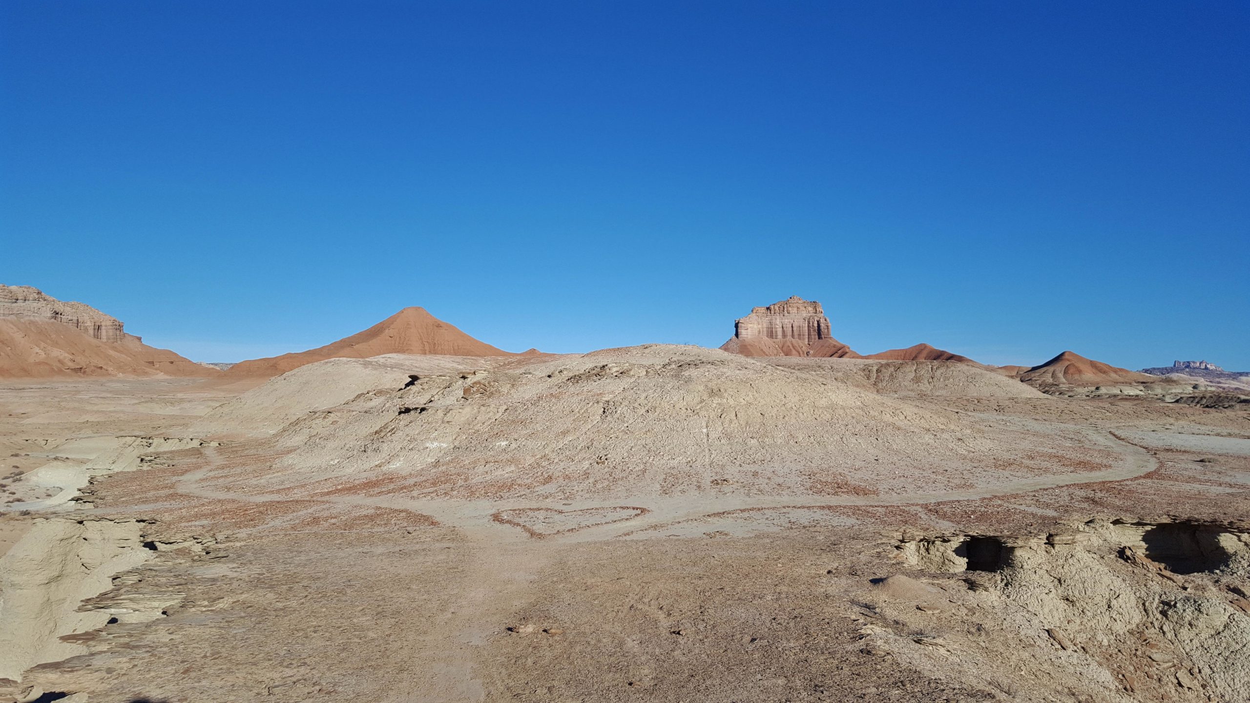 A barren landscape featuring various reddish hills and rock formations under a clear blue sky. The ground shows a heart shape carved into the earth, surrounded by dry, textured terrain. Goblin Valley State Park mountain bike trail.