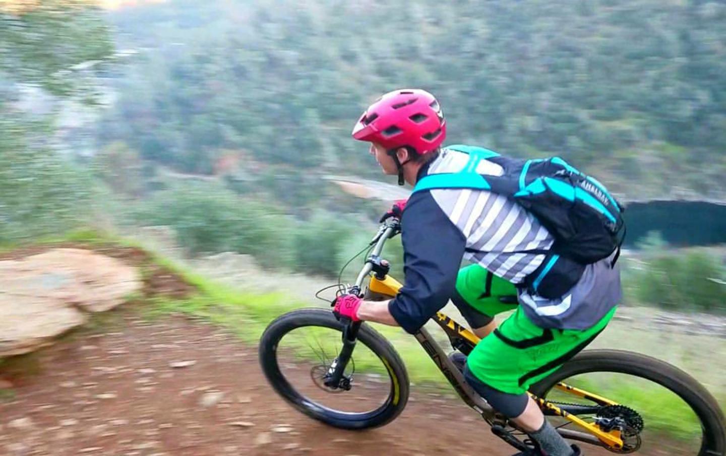 A mountain biker is riding downhill on a rocky trail, leaning into the turn. The cyclist is wearing a red helmet, a striped shirt, and bright green shorts. A backpack is secured on their back, and the background features a scenic view of greenery and a body of water below. The motion blur conveys speed and excitement. Stagecoach / Flood / Manzanita mountain bike trail.