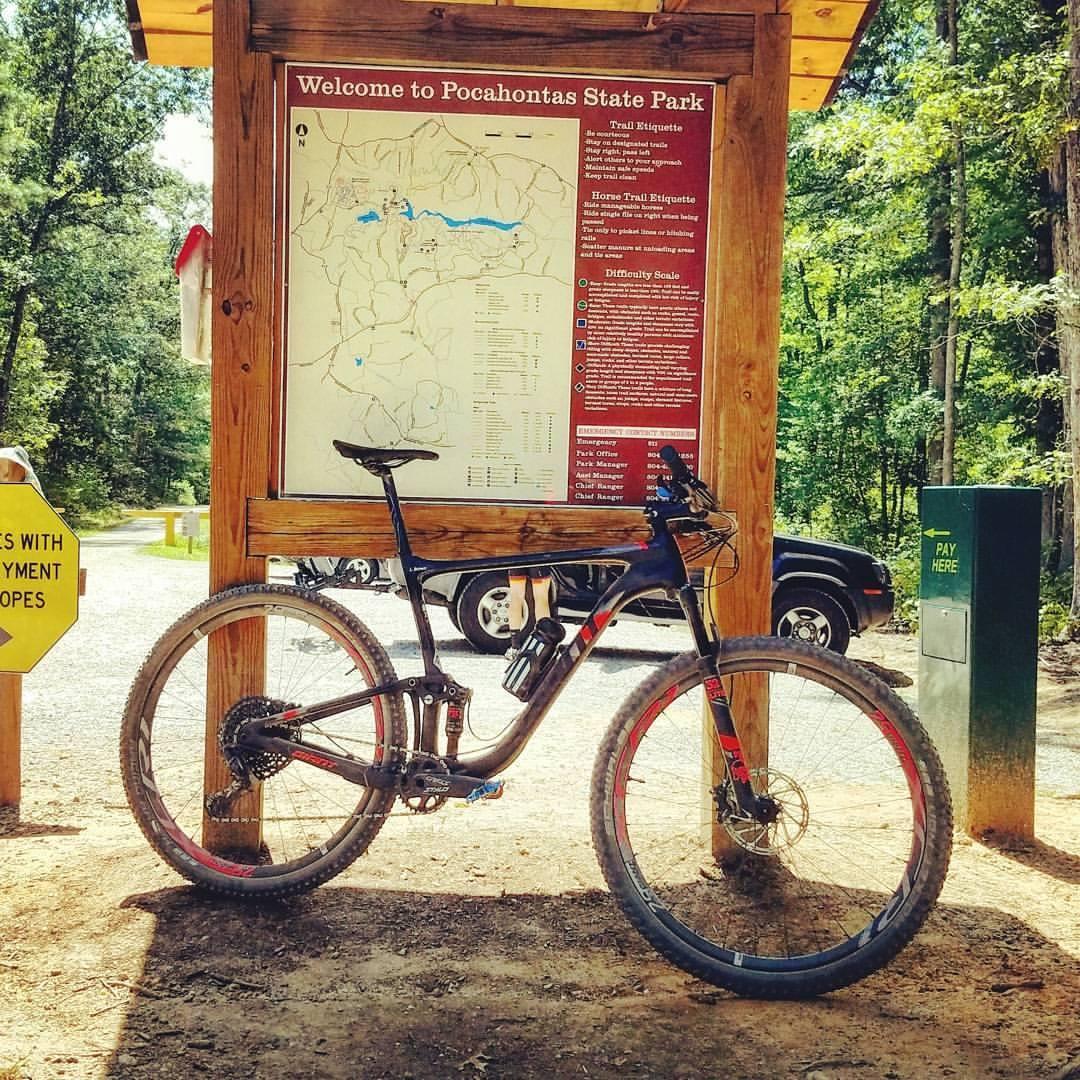 Giant Anthem Advanced Pro 29: A mountain bike leaning against a wooden sign at the entrance of Pocahontas State Park. The sign displays a map of the park along with trail etiquette information. In the background, a pay station and a vehicle are visible amidst a wooded setting.