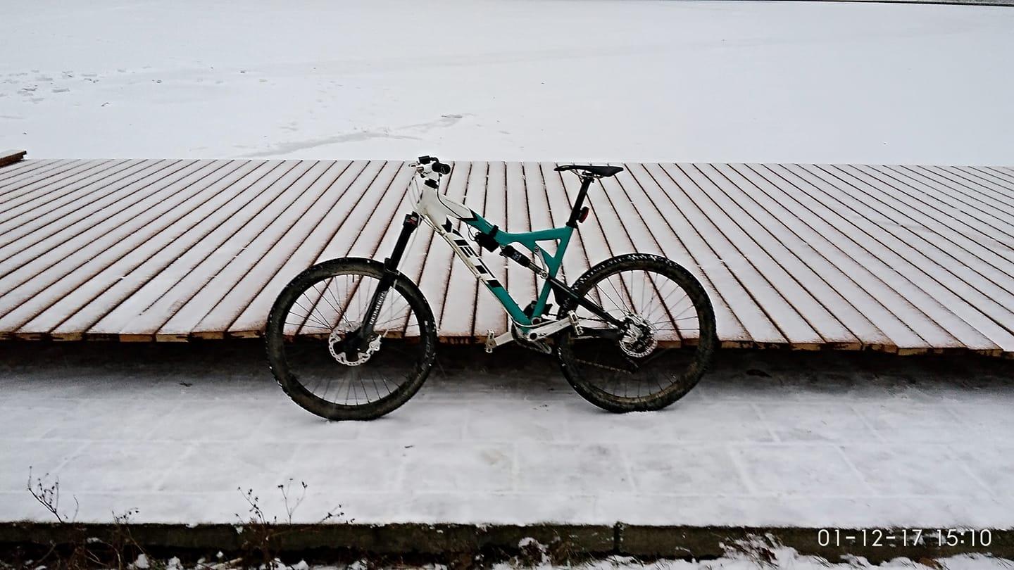Yeti 575: A mountain bike with a white and turquoise frame is parked on a wooden platform covered with snow. The background features a frozen surface, creating a winter setting, with soft, diffused lighting.
