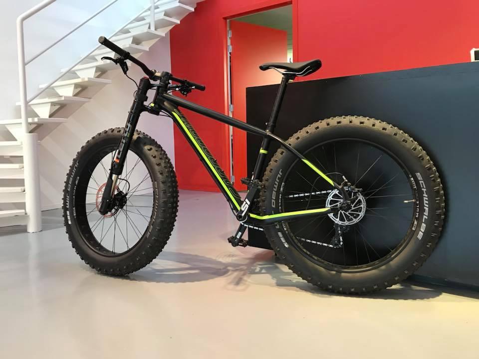 Cannondale Fat CAAD 1: A black fat tire bike with neon green accents stands on a light gray floor. The bike is positioned next to a black display counter, with a staircase and a doorway in the background, featuring red walls.