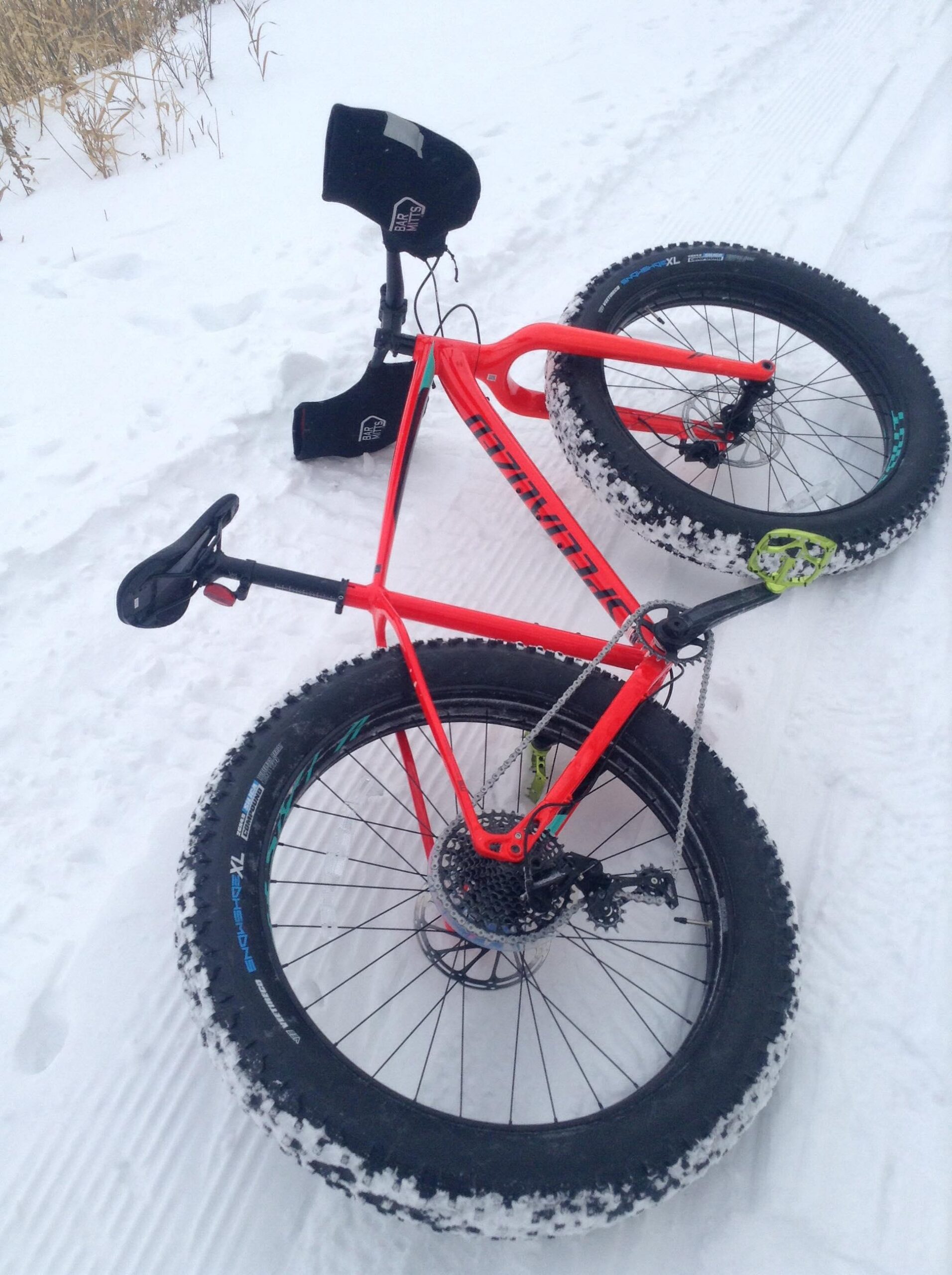 Specialized Fatboy: A bright orange fat bike rests on a snow-covered surface, with thick tires showing signs of snow accumulation. The bike features a streamlined frame, a black saddle, and a chain system. Nearby, a snow guard is positioned, indicating winter cycling readiness. The background includes a patch of snowy terrain and some blurred grass.