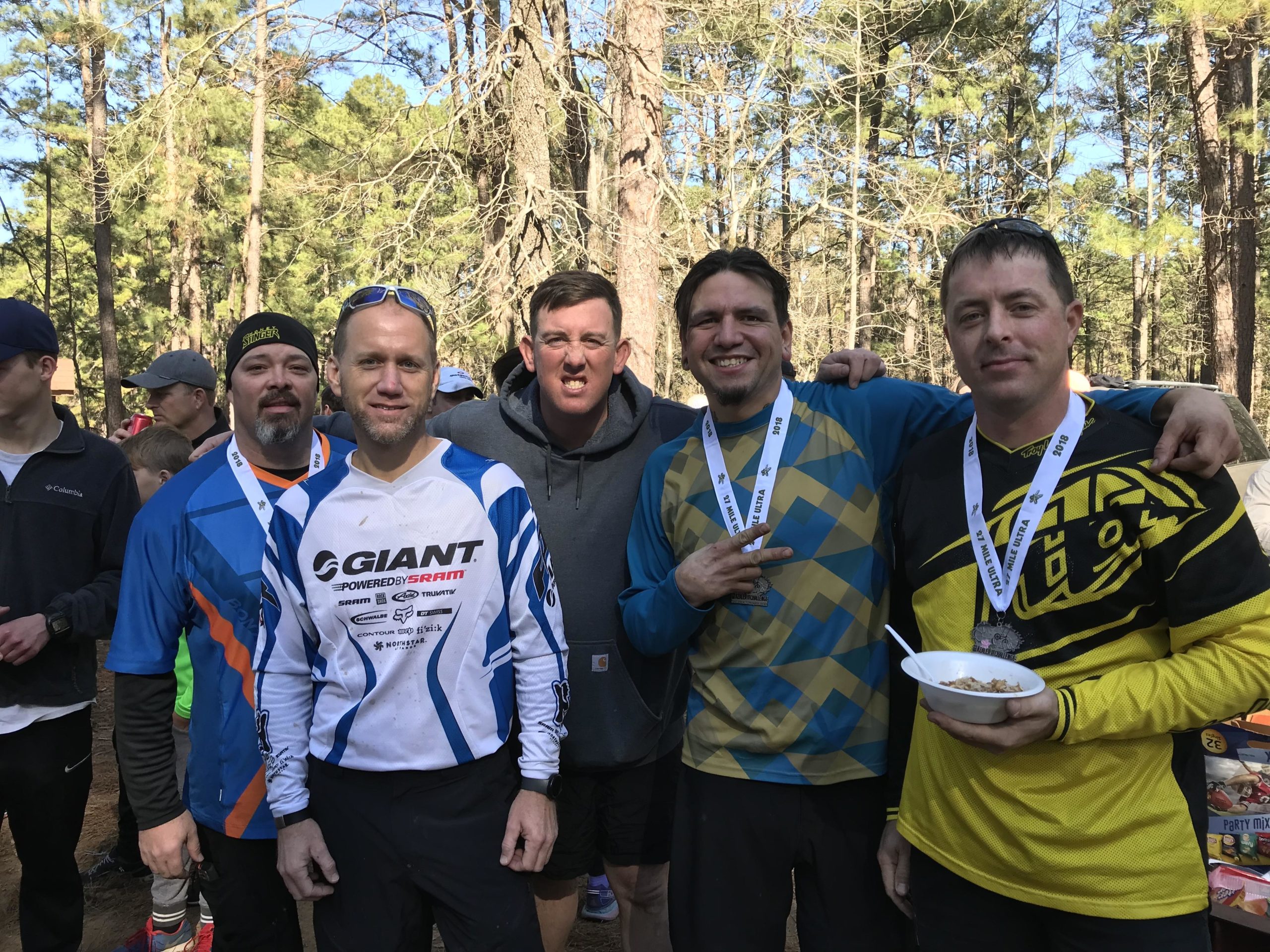 Five men stand together in a wooded area, smiling and posing for the camera. They are wearing colorful jerseys and medals around their necks, indicating participation in a biking event. One man is holding a bowl of food. The background features tall trees and a crowd of people nearby. Wild Azalea Trail mountain bike trail.