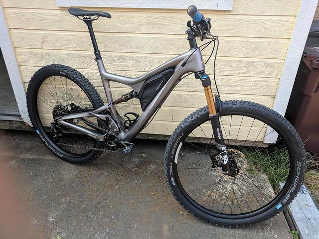 Ibis Ripley 29: A modern mountain bike with a sleek gray frame, featuring front suspension and wide tires, positioned beside a yellow wooden wall and a dark trash bin. The bike is angled to showcase its components, including the handlebars and drivetrain.