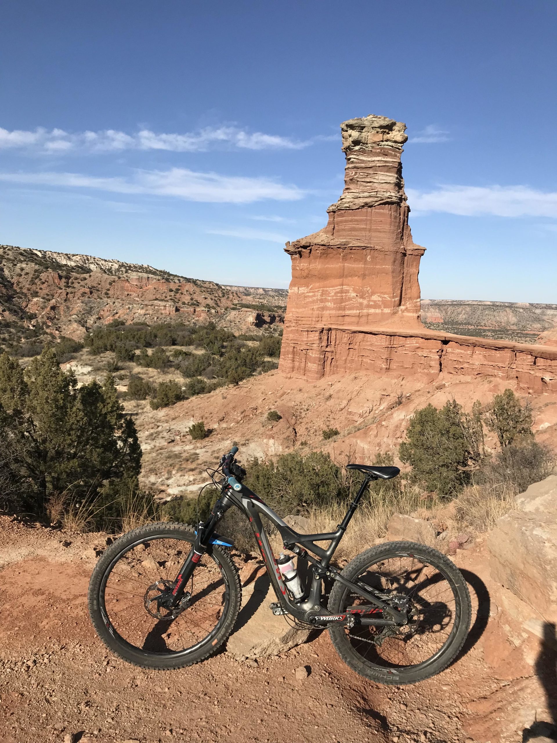 A mountain bike resting on a rocky trail with a prominent rock formation in the background, set against a clear blue sky. The landscape features red and brown tones, typical of a canyon environment, with sparse vegetation. Palo Duro Canyon mountain bike trail.