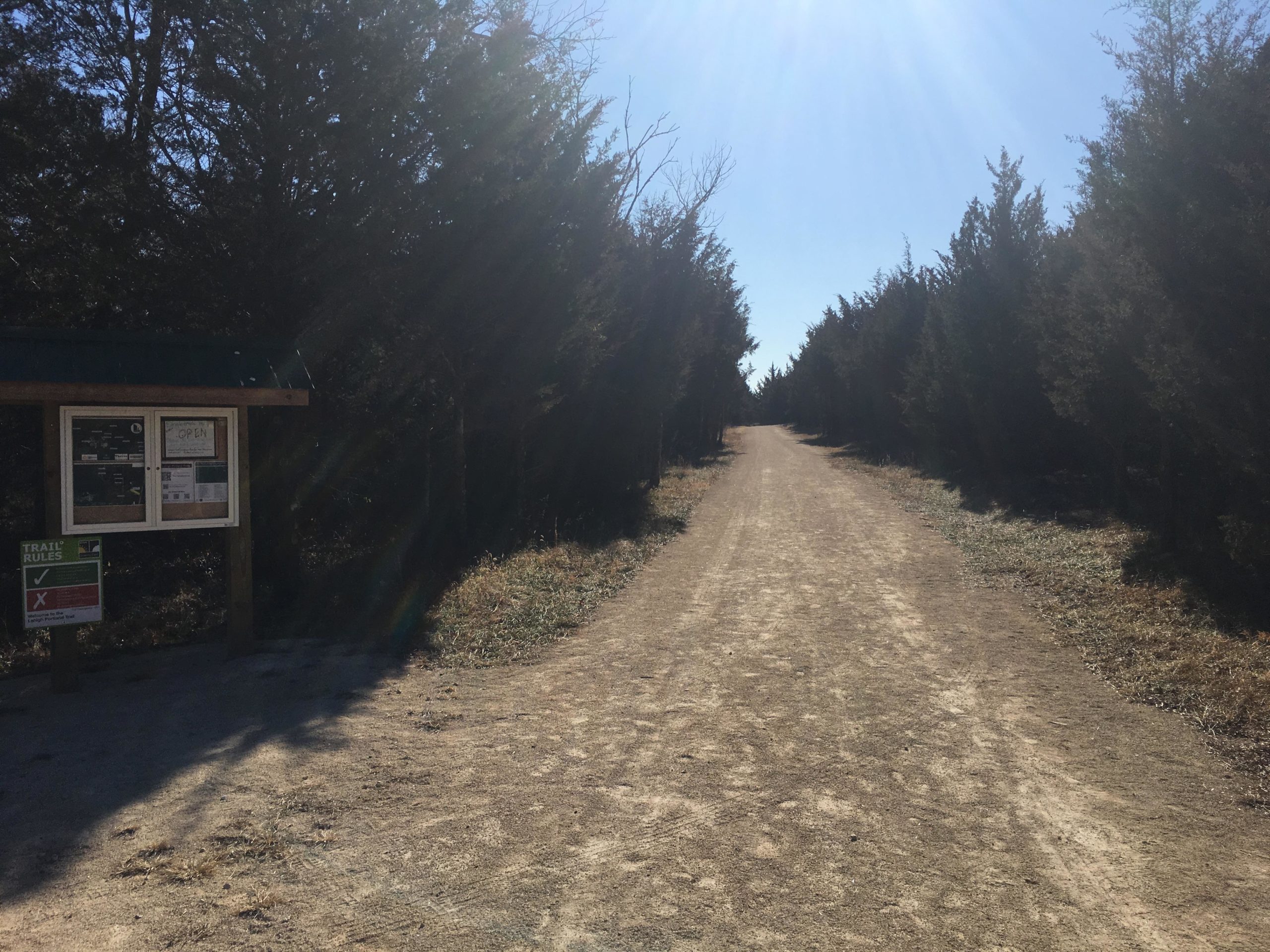 A dirt path lined with trees leads into the distance, with a trail information sign visible on the left side. The sign displays trail rules and indicates that the area is open. The scene is bright and sunny, with clear blue skies above. Lehigh Portland Trails mountain bike trail.