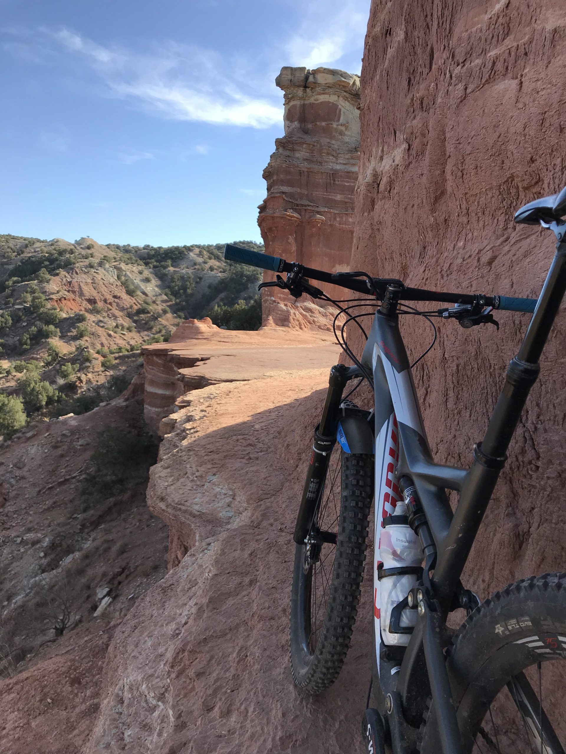 Alt text: A mountain bike is resting on a rocky ledge overlooking a canyon landscape under a clear blue sky. The terrain features steep, reddish cliffs and lush greenery in the distance. Palo Duro Canyon mountain bike trail.
