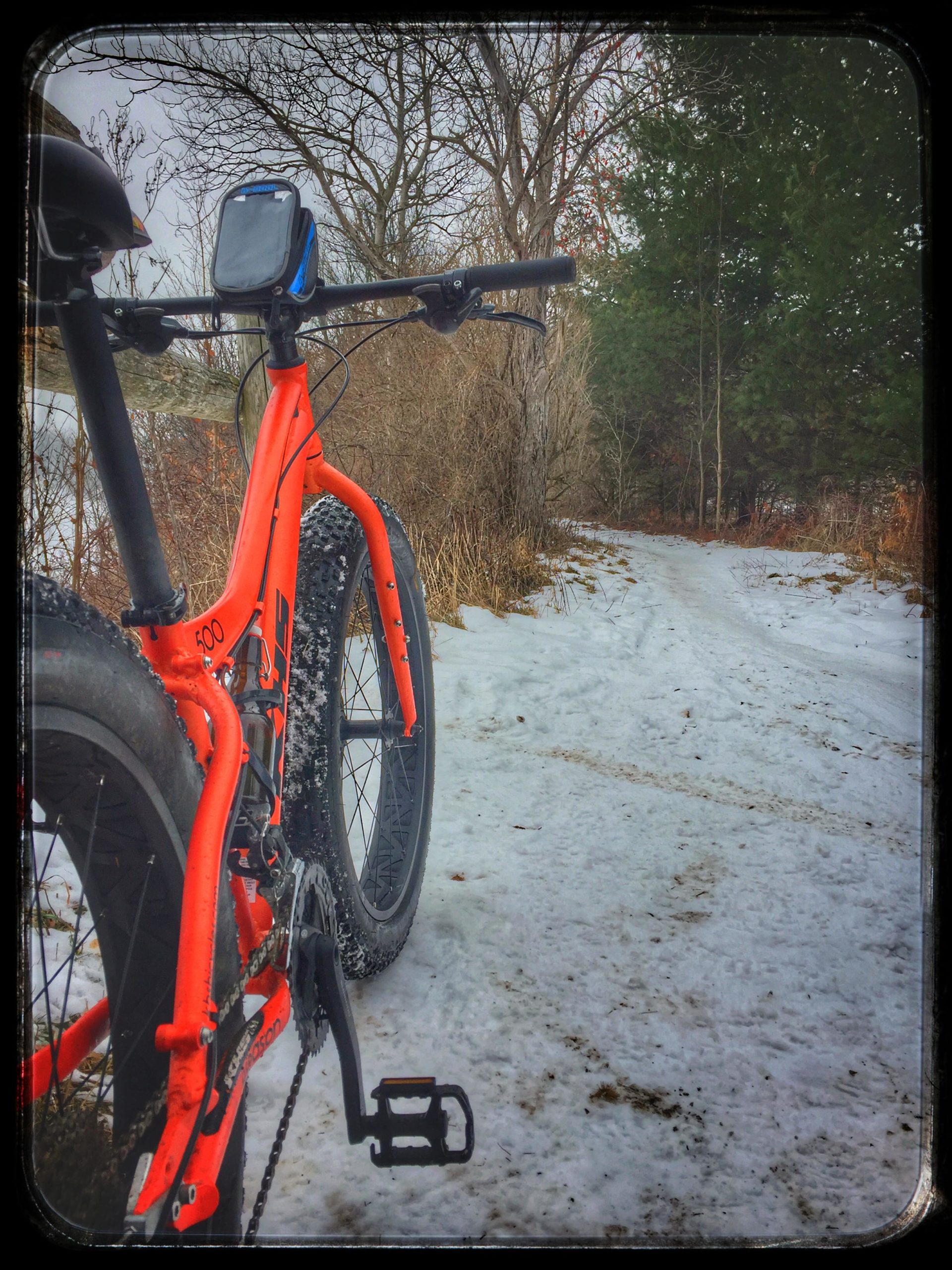 A close-up view of an orange fat bike parked on a snow-covered trail. The bike’s handlebars, equipped with a GPS device, are prominent in the foreground, while a winding path surrounded by trees and brush leads into the distance. The scene is set in a wintry landscape. Fanshawe Lake mountain bike trail.