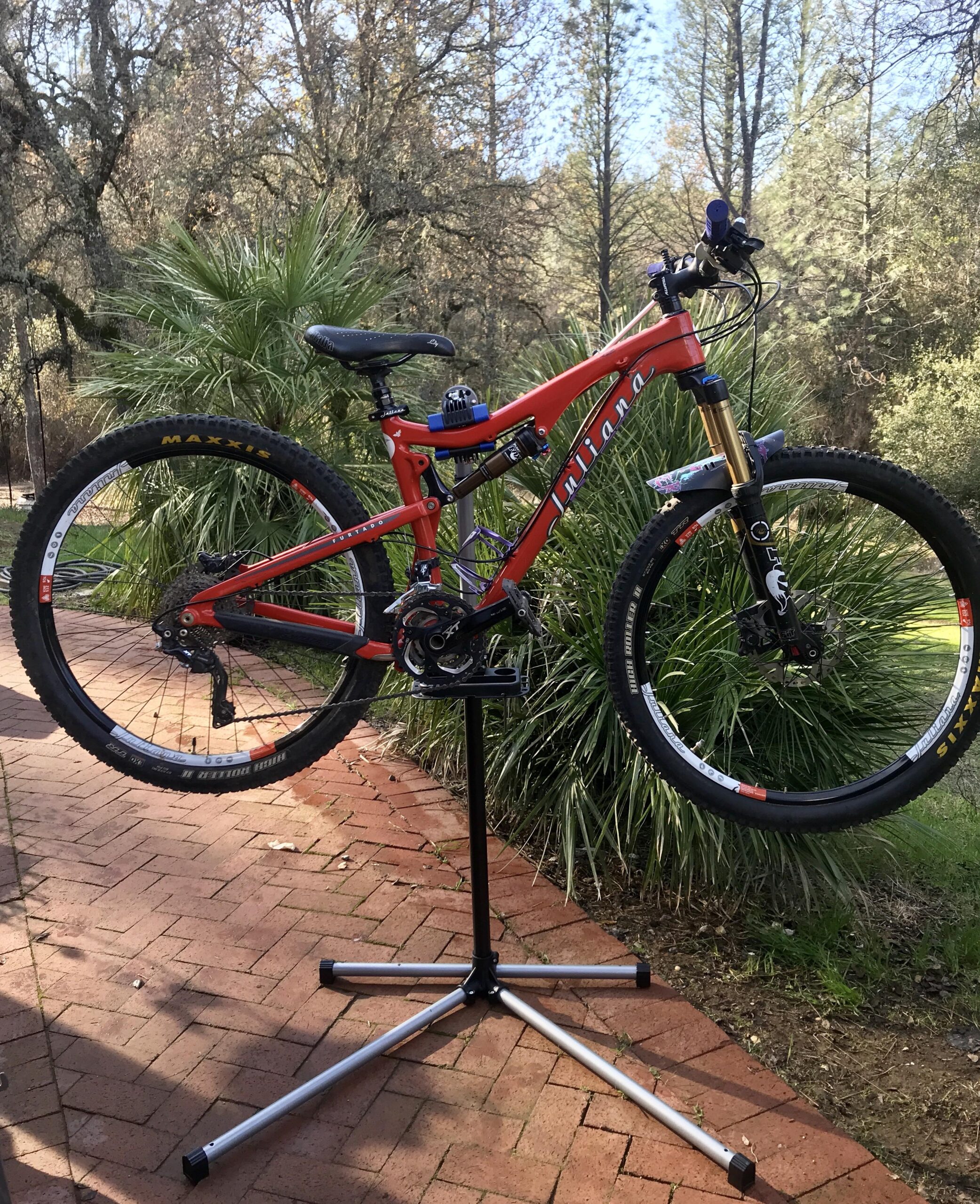 Juliana Furtado: A vibrant red mountain bike is positioned upright on a bike repair stand, set against a backdrop of lush greenery and trees. The bike features thick tires designed for off-road traction and is standing on a brick pathway.