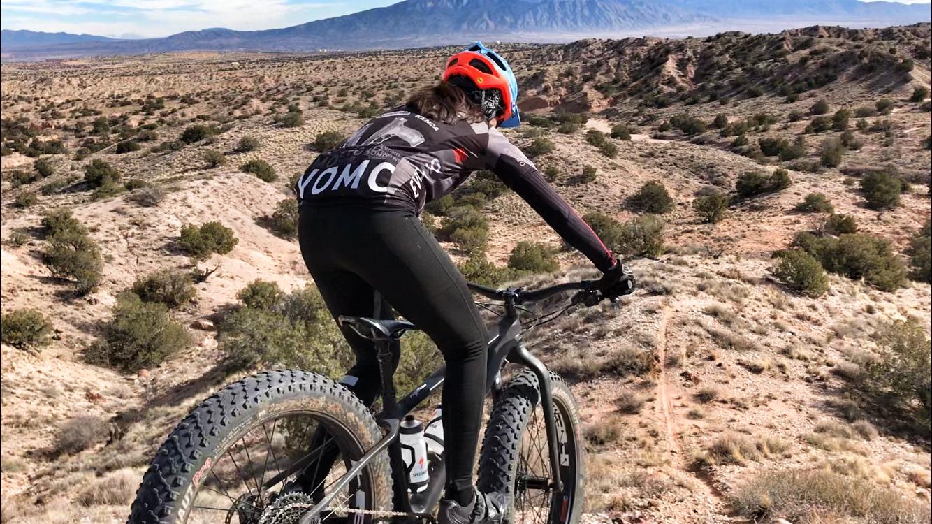 A cyclist wearing a red helmet and a black jersey with "YOMC" printed on it is riding a mountain bike on a rocky trail. In the background, expansive desert terrain is visible, with rolling hills and sparse vegetation under a clear blue sky. Mariposa Fat Bike Trails mountain bike trail.