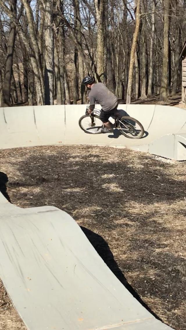 A person wearing a helmet rides a bike around a concrete pump track in a wooded area. The track features smooth, curved walls and is surrounded by bare trees and patches of dry grass. Two Rivers Bike Park mountain bike trail.