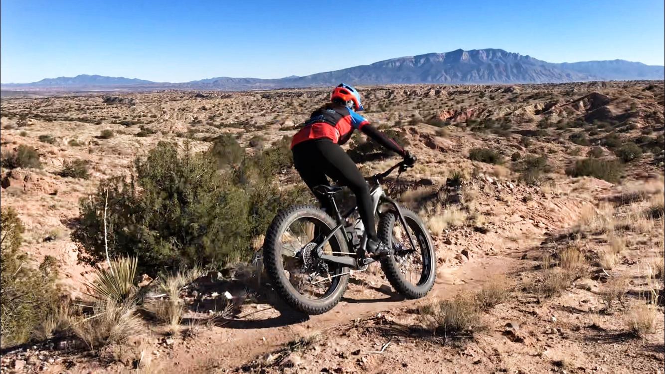A person riding a fat tire mountain bike along a dirt trail in a rocky desert landscape, with mountains in the background and a clear blue sky. Mariposa Fat Bike Trails mountain bike trail.