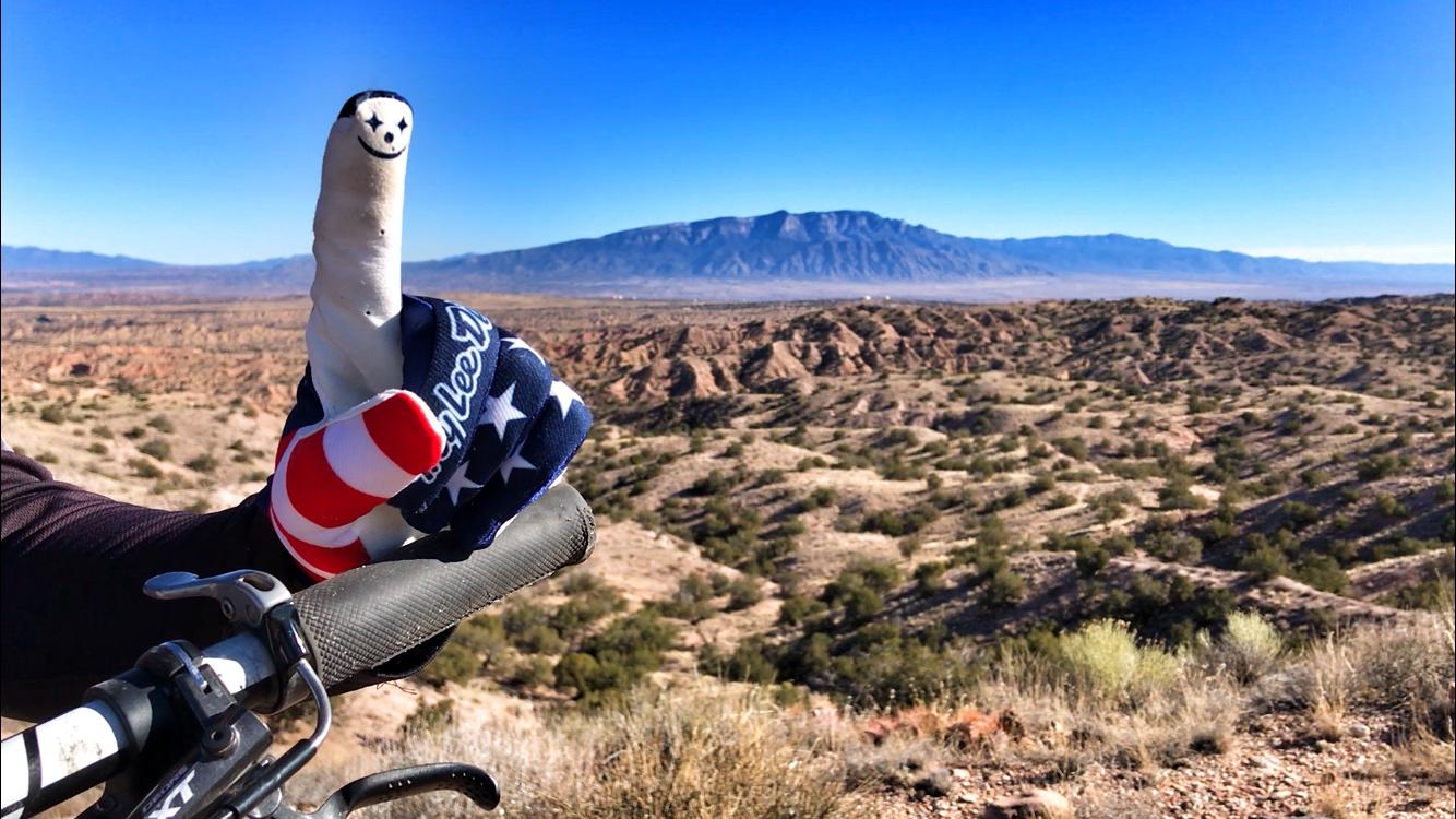 A mountain landscape under a clear blue sky, with a close-up of a hand wearing a patriotic-themed glove holding a bike handlebar and displaying a finger with a smiley face. The terrain features rugged hills and sparse vegetation, creating a scenic backdrop. Mariposa Fat Bike Trails mountain bike trail.
