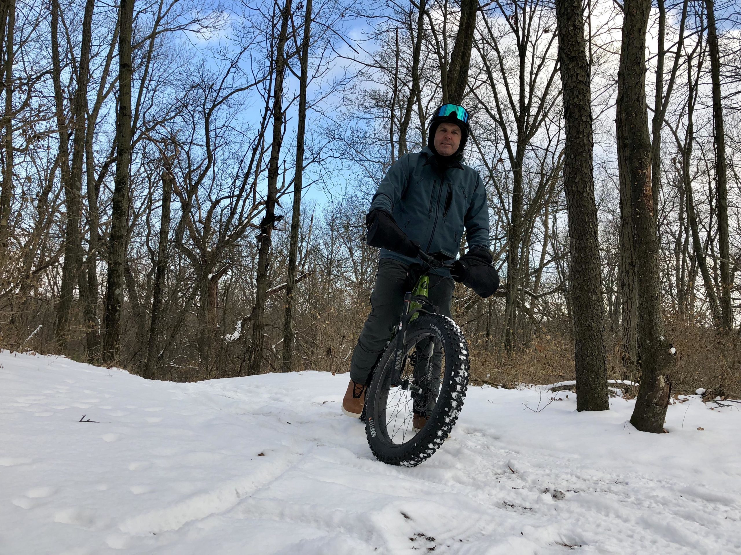 A person riding a fat bike on a snow-covered trail in a forest, surrounded by bare trees. The cyclist is wearing a helmet and warm clothing, with their hands covered by mittens. The sky is partly cloudy. Kettle Moraine John Muir + Emma Carlin mountain bike trail.