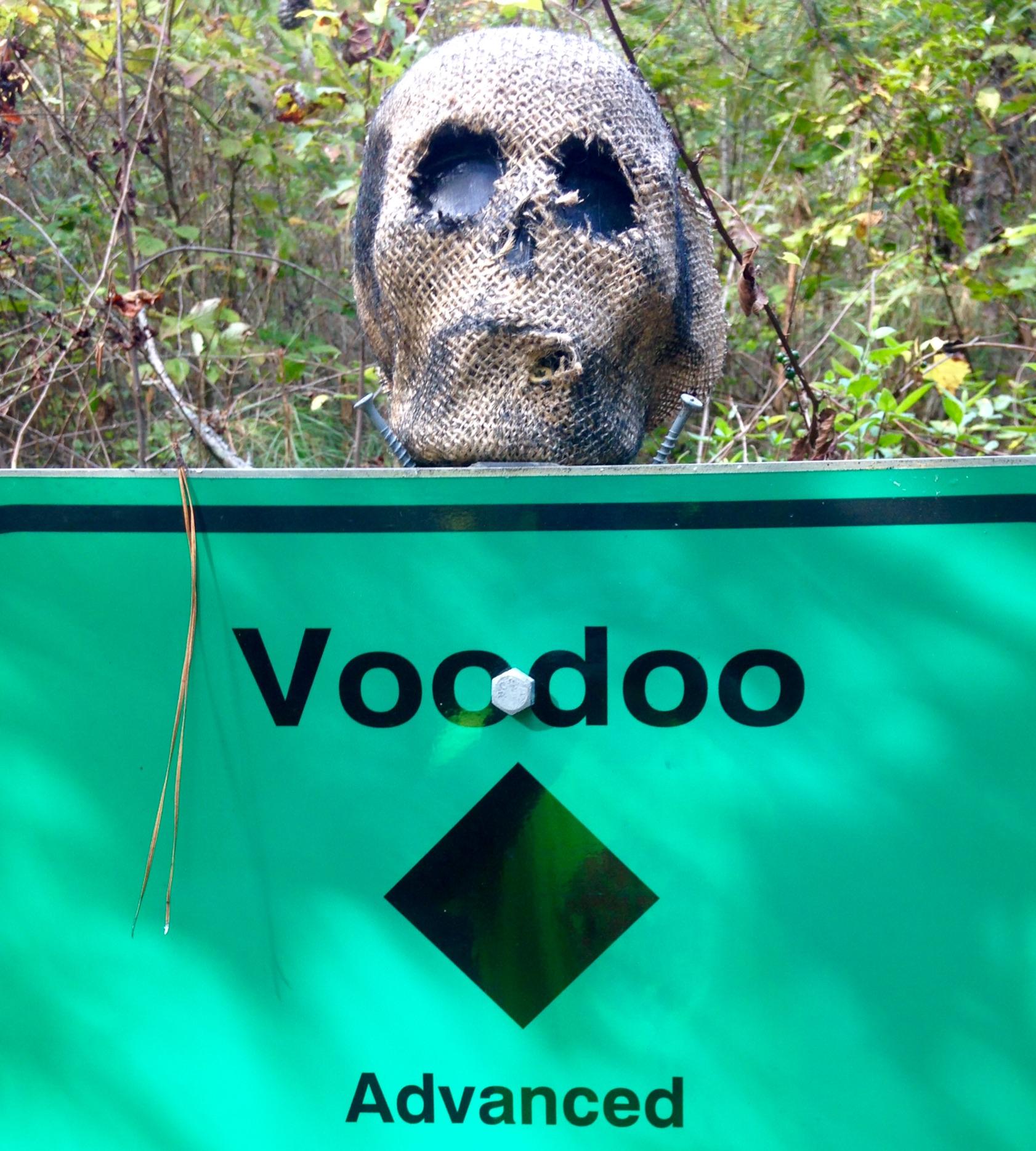 A burlap skull is perched atop a green sign that reads "Voodoo" in bold letters, with the word "Advanced" below it, accompanied by a black diamond symbol. The background features greenery, suggesting a forested area. Allatoona Creek Park mountain bike trail.