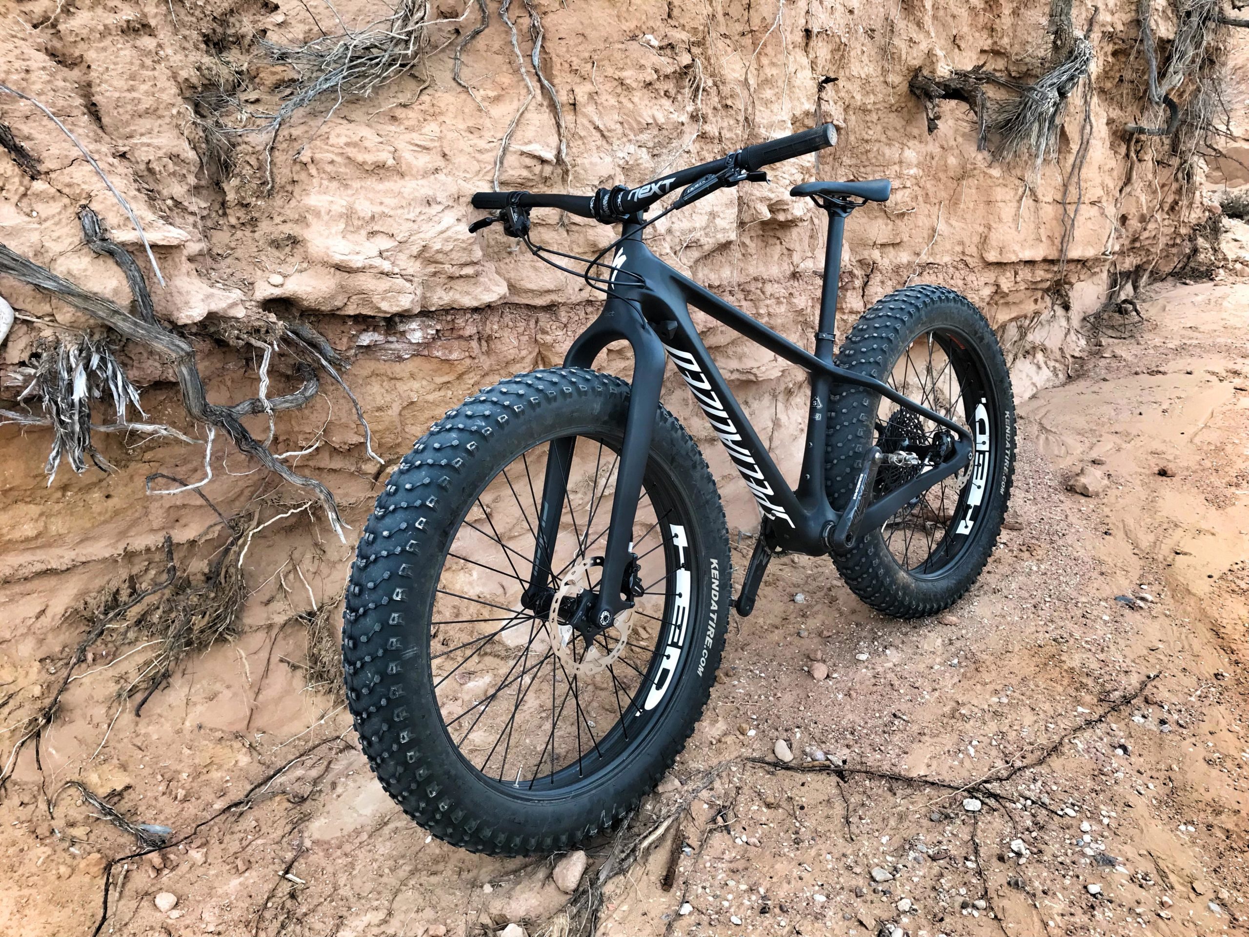 A black fat tire mountain bike is leaning against a rocky, sandy terrain with exposed roots and a textured earth background. The bike features wide, knobby tires designed for off-road riding, and has a sleek, aerodynamic frame. Mariposa Fat Bike Trails mountain bike trail.