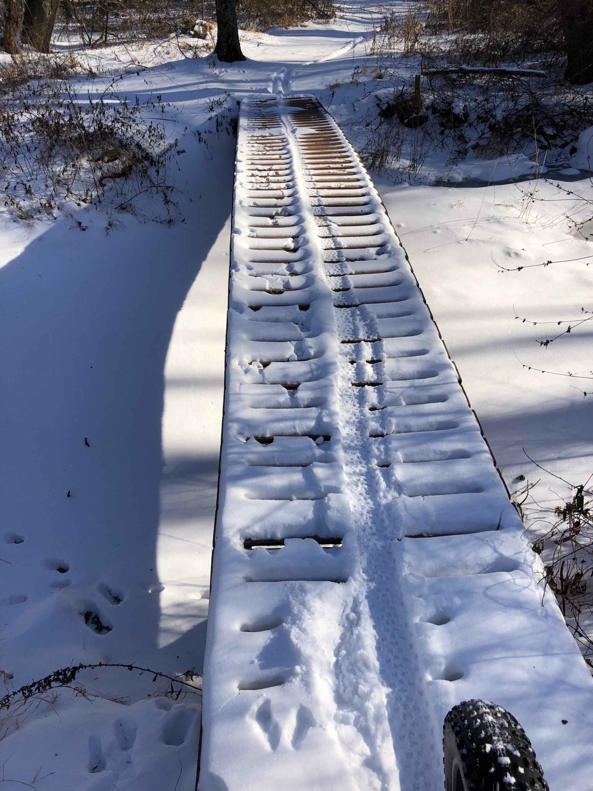 A snow-covered wooden bridge in a forest, with visible footprints and tire tracks in the snow, leading towards a path lined by trees. The scene is bright and wintery, showcasing a tranquil outdoor environment. Six Mile Run mountain bike trail.