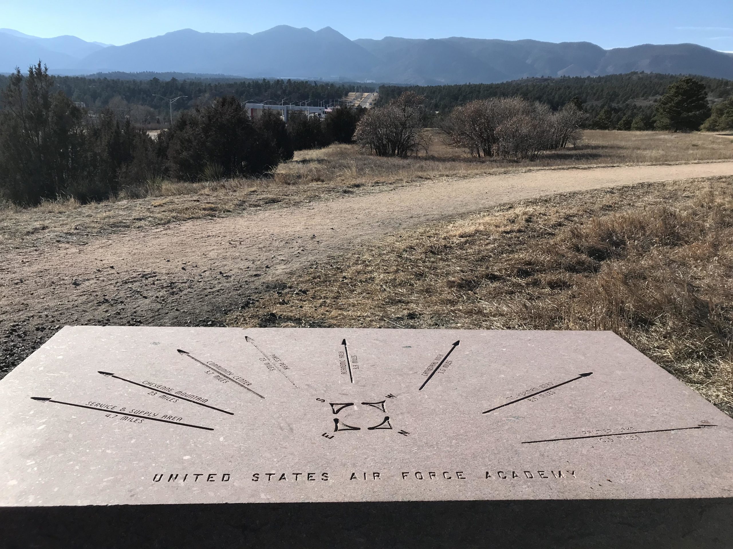 A stone marker with directional indicators engraved on its surface, showing distances to various landmarks, set against a backdrop of mountains and a clear blue sky. The scene includes a dirt path leading away from the marker, with sparse vegetation and trees in the surrounding area. The marker is labeled "United States Air Force Academy." Santa Fe Trail mountain bike trail.