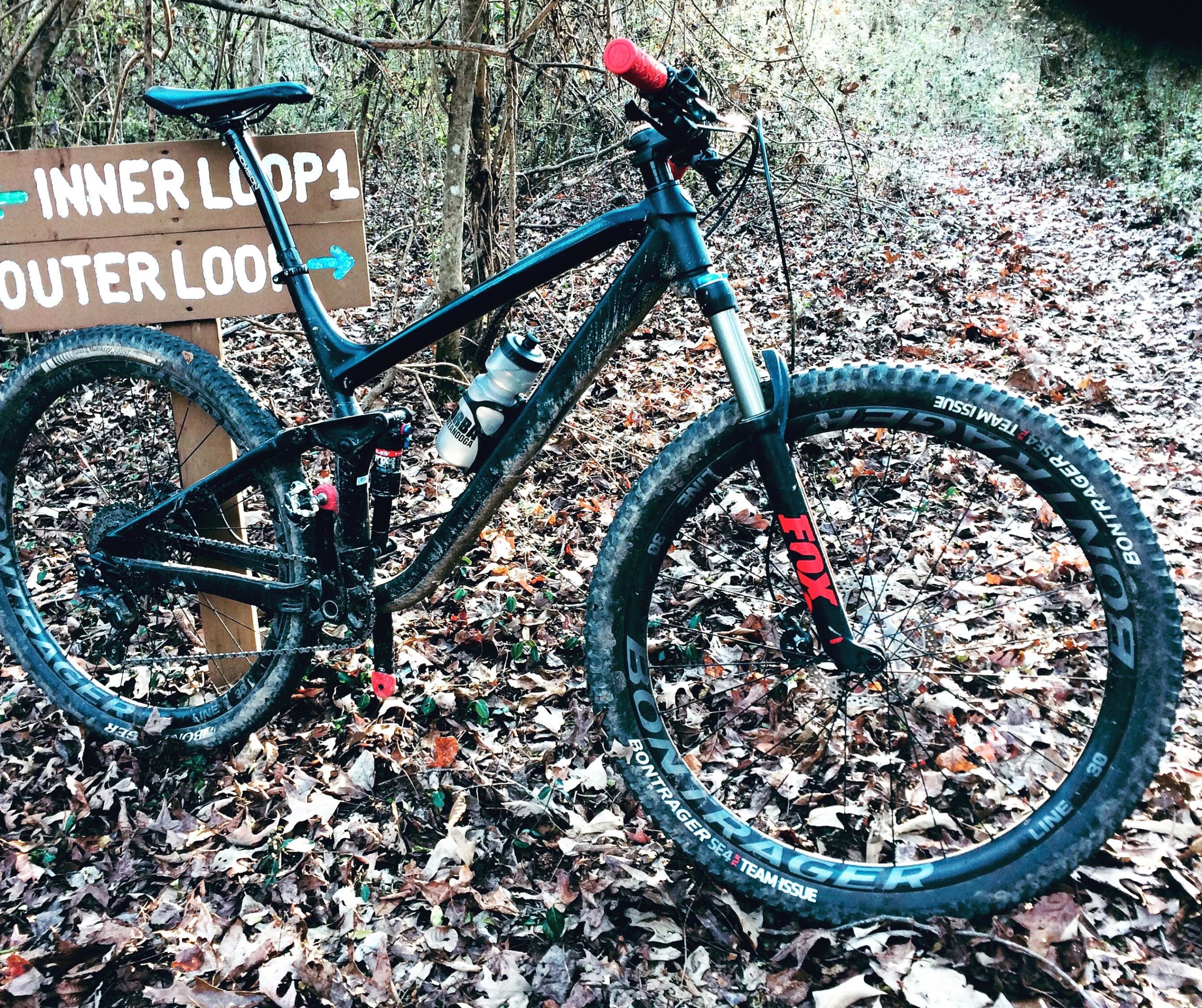 A mountain bike parked next to a wooden trail sign that reads "INNER LOOP 1" and "OUTER LOOP" with arrows indicating directions. The bike features a black frame, red handle grips, and is positioned on a forested trail covered in fallen leaves. Booker T. Washington State Park mountain bike trail.