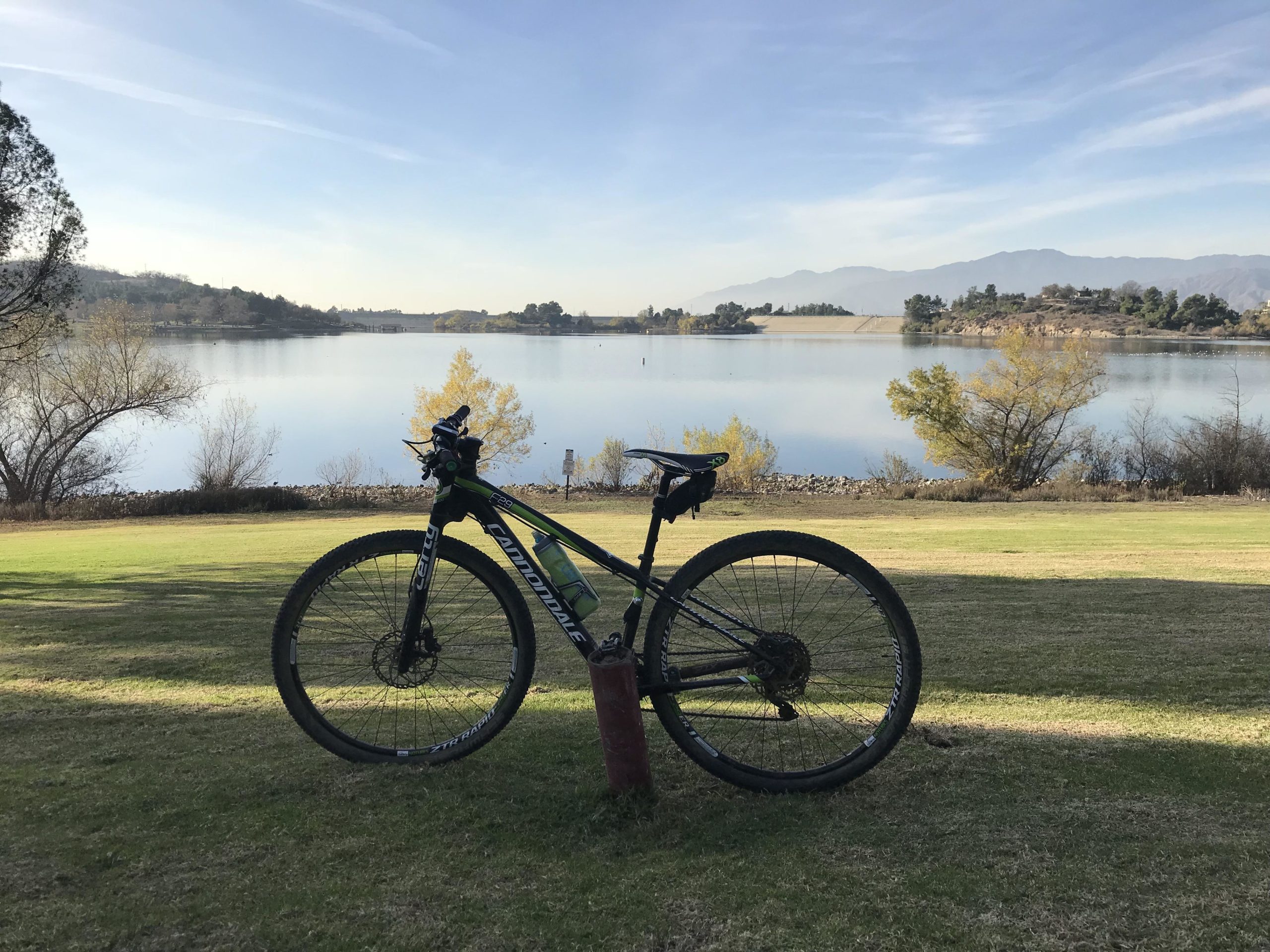 A mountain bike rests on its kickstand in a grassy area, overlooking a calm lake surrounded by trees and distant hills. The sky is clear with soft clouds, and a few patches of autumn foliage can be seen nearby. Frank G. Bonelli Regional Park mountain bike trail.