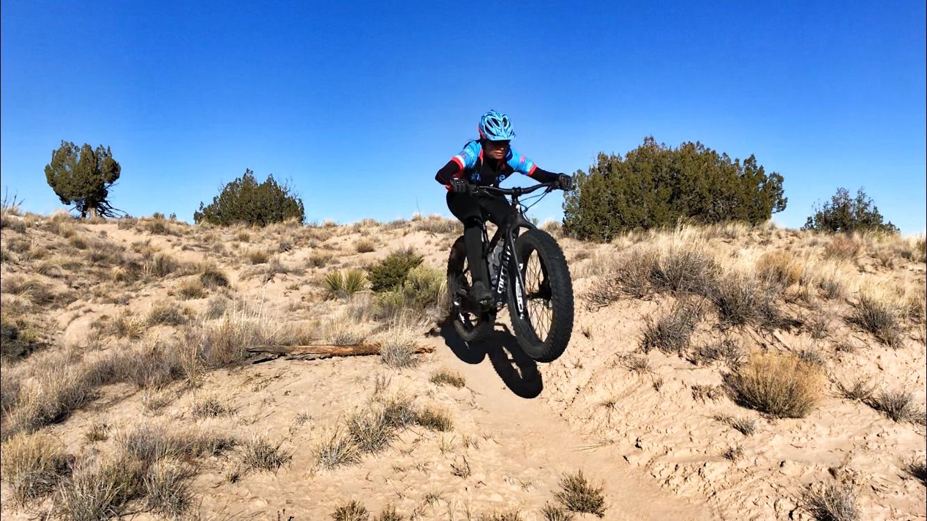 A person in cycling gear rides a fat bike over a sandy trail, jumping off a small incline against a backdrop of blue sky and sparse vegetation. Mariposa Fat Bike Trails mountain bike trail.