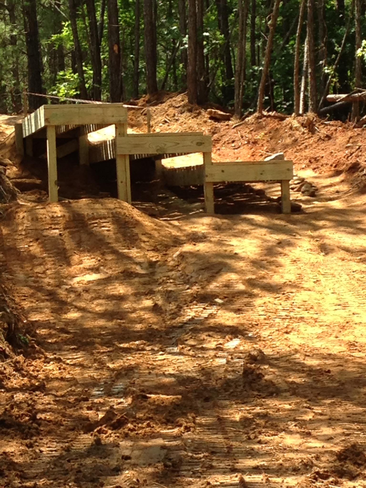 A wooden ramp with multiple levels, set against a backdrop of trees and a dirt path. The ramp appears to be in a forested area, with the ground surrounding it showing signs of recent construction or excavation. Sunlight filters through the trees, casting shadows on the ground. Allatoona Creek Park mountain bike trail.