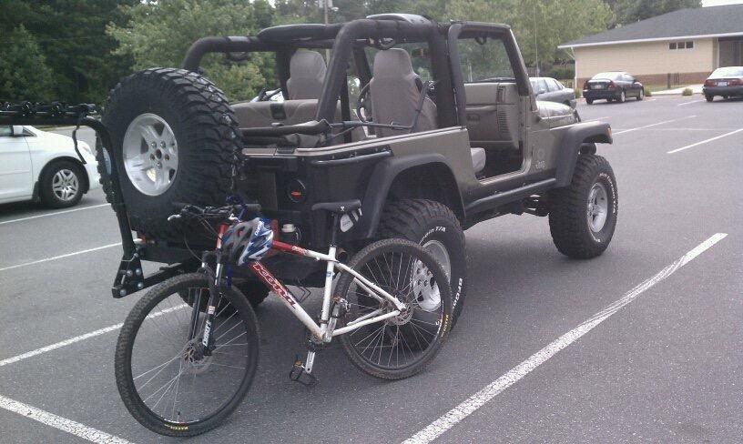 Kona Kahuna: A sporty off-road vehicle parked in a lot with its rear-mounted spare tire visible, alongside a mountain bike secured to the vehicle's frame. The scene captures a mix of outdoor adventure and practical transport.