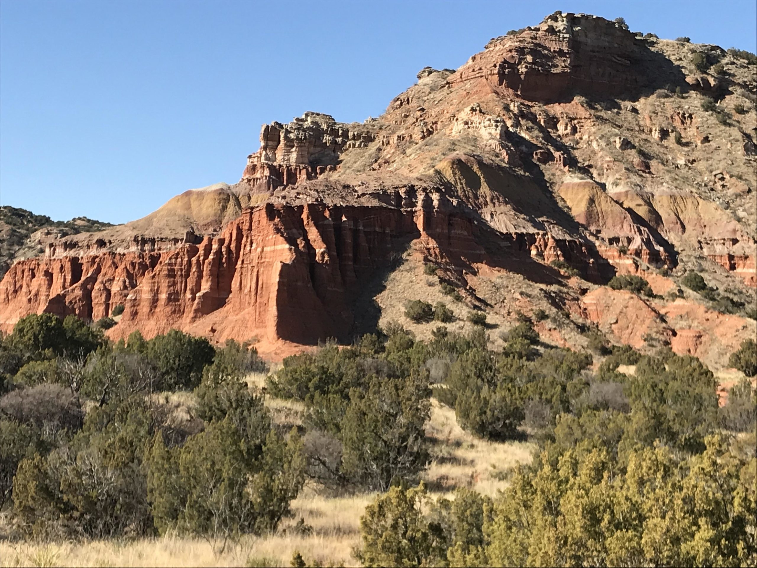 A scenic view of rugged reddish-brown cliffs and hills under a clear blue sky, with patches of green vegetation in the foreground and a gentle slope leading up to the rocky formations. Palo Duro Canyon mountain bike trail.
