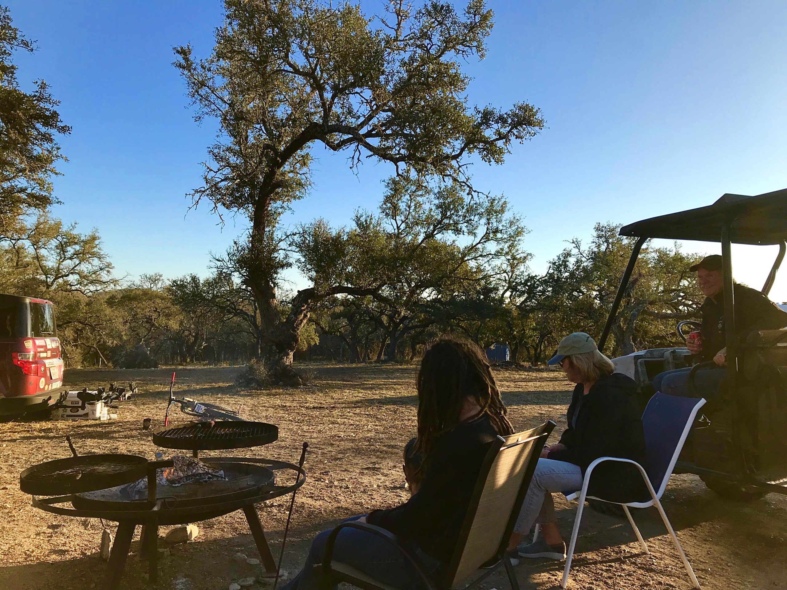 A group of people sitting around a fire pit in a natural outdoor setting, with a blue sky overhead. Some individuals are seated on camping chairs, while one person is sitting in a golf cart, holding a drink. The area is surrounded by trees, and a vehicle is parked nearby. The scene captures a relaxed atmosphere, suggesting a gathering or camping experience. Flat Rock Ranch mountain bike trail.