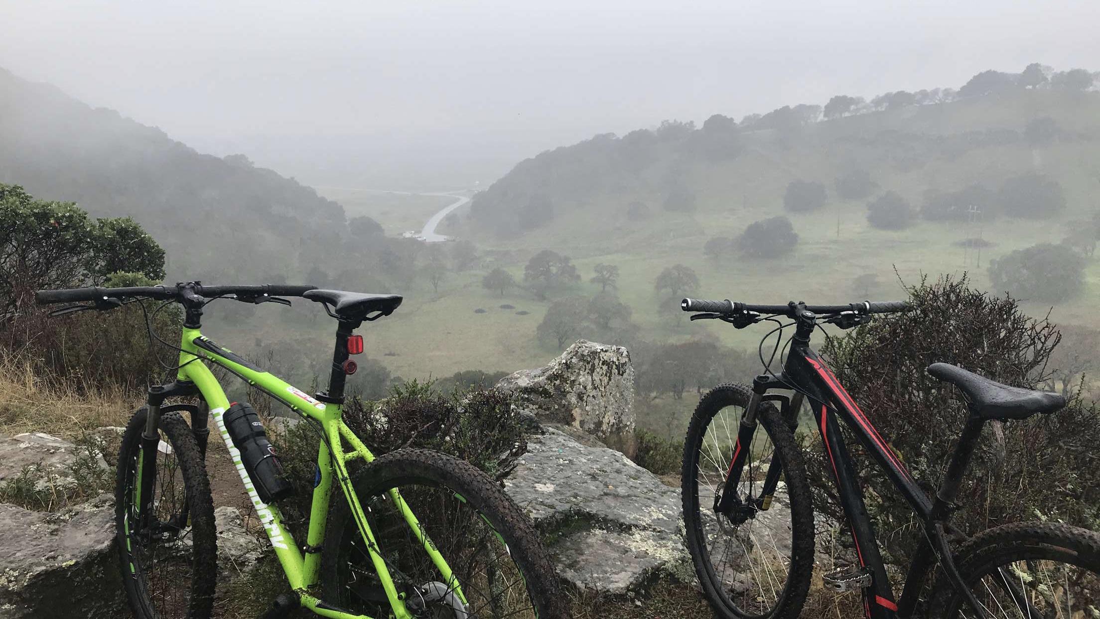 A scenic view from a rocky overlook featuring two mountain bikes, one green and one black, positioned side by side. In the background, a misty landscape with rolling hills and a winding road can be seen, shrouded in fog. The area is surrounded by shrubs and grass, conveying a sense of adventure in nature. Rockville Park mountain bike trail.