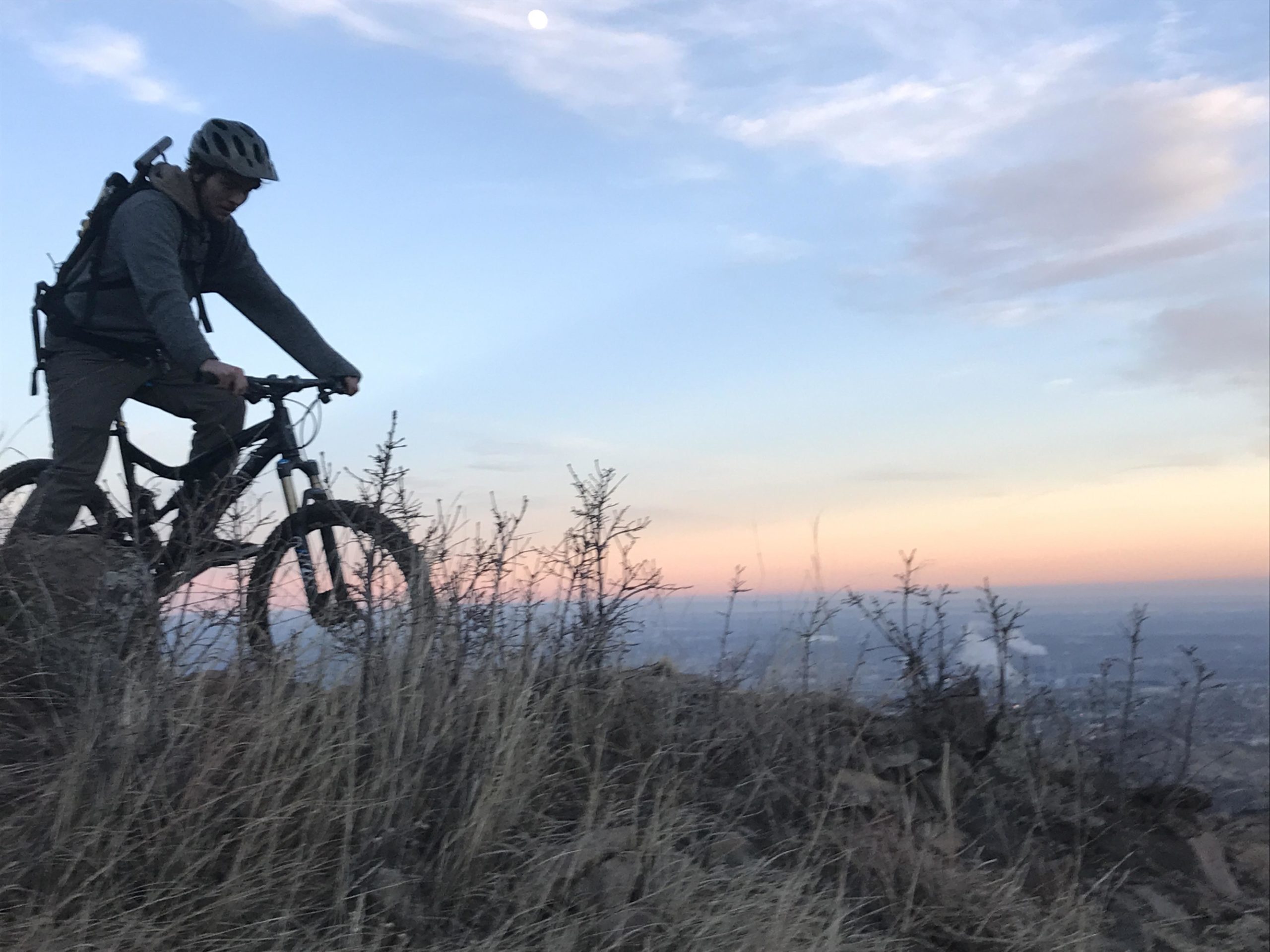 A mountain biker navigating a rocky terrain at dusk, with a scenic background of a pastel sky and distant city lights. The biker is wearing a helmet and a backpack, and is surrounded by tall grasses and sparse shrubs. Red Rock Canyon mountain bike trail.