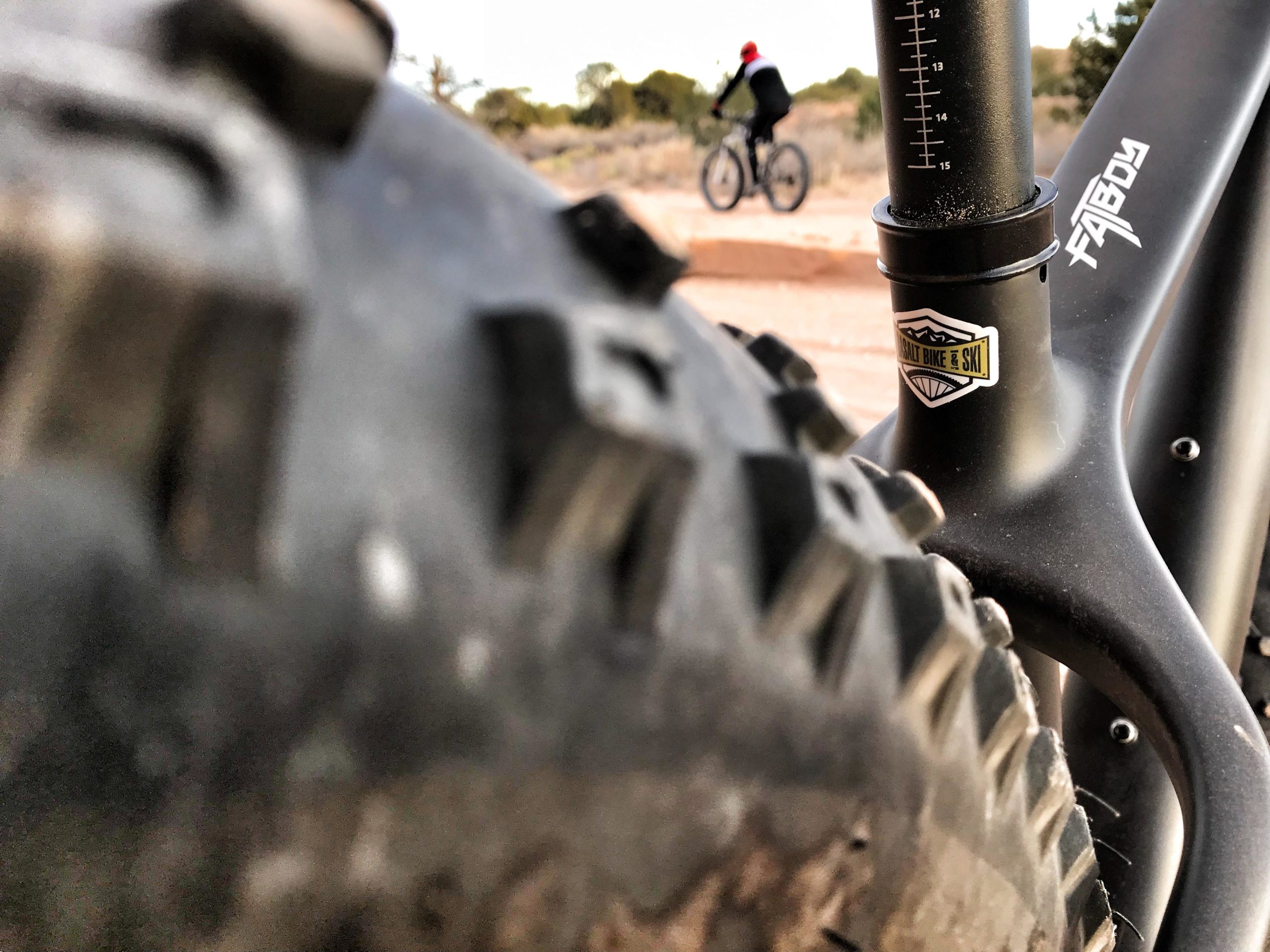 A close-up view of a mountain bike tire and frame, showcasing the tread patterns and a bike shop sticker. In the background, a blurred figure is riding another bike on a gravel path surrounded by natural scenery. Mariposa Fat Bike Trails mountain bike trail.