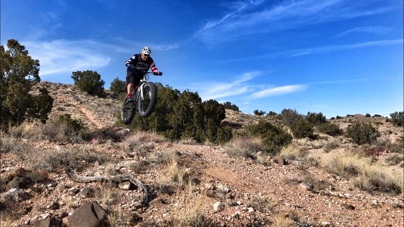 A mountain biker performing a jump over a rocky terrain, surrounded by shrubs and a clear blue sky. Mariposa Fat Bike Trails mountain bike trail.
