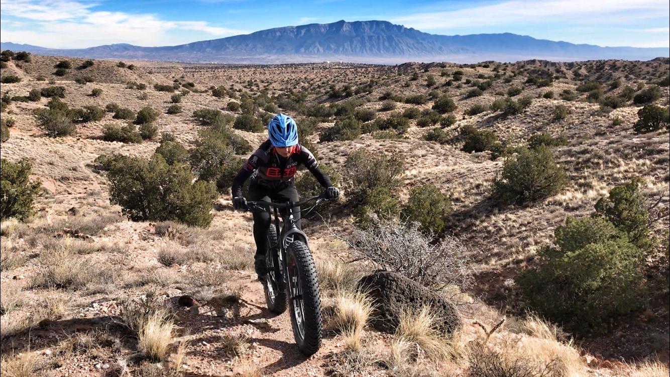 A person riding a fat bike on a rugged, rocky trail in a desert landscape, with sparse vegetation and mountains in the background under a clear blue sky. Mariposa Fat Bike Trails mountain bike trail.