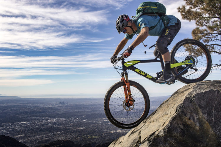 A mountain biker in a blue shirt and helmet navigates a rocky ledge, leaning forward on a brightly colored bike with orange and green accents. The background features a panoramic view of a city skyline, surrounded by a vast landscape and a clear blue sky with wispy clouds.