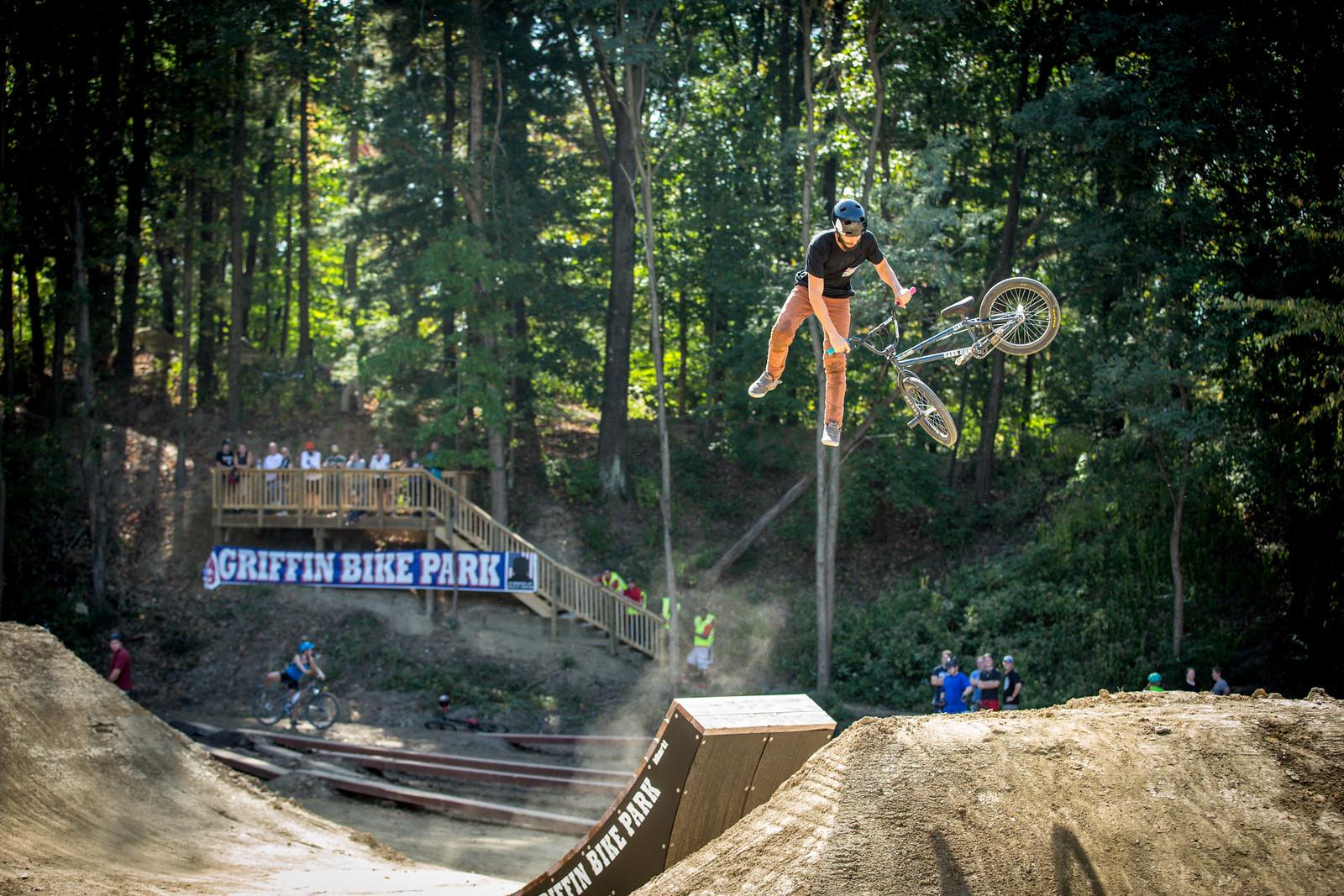 A BMX rider performing a trick mid-air above a dirt ramp at Griffin Bike Park, surrounded by trees. Spectators watch from a wooden platform in the background. Dust is kicked up from the ramp as the rider, wearing a helmet and casual attire, showcases their skills. Griffin Bike Park mountain bike trail.