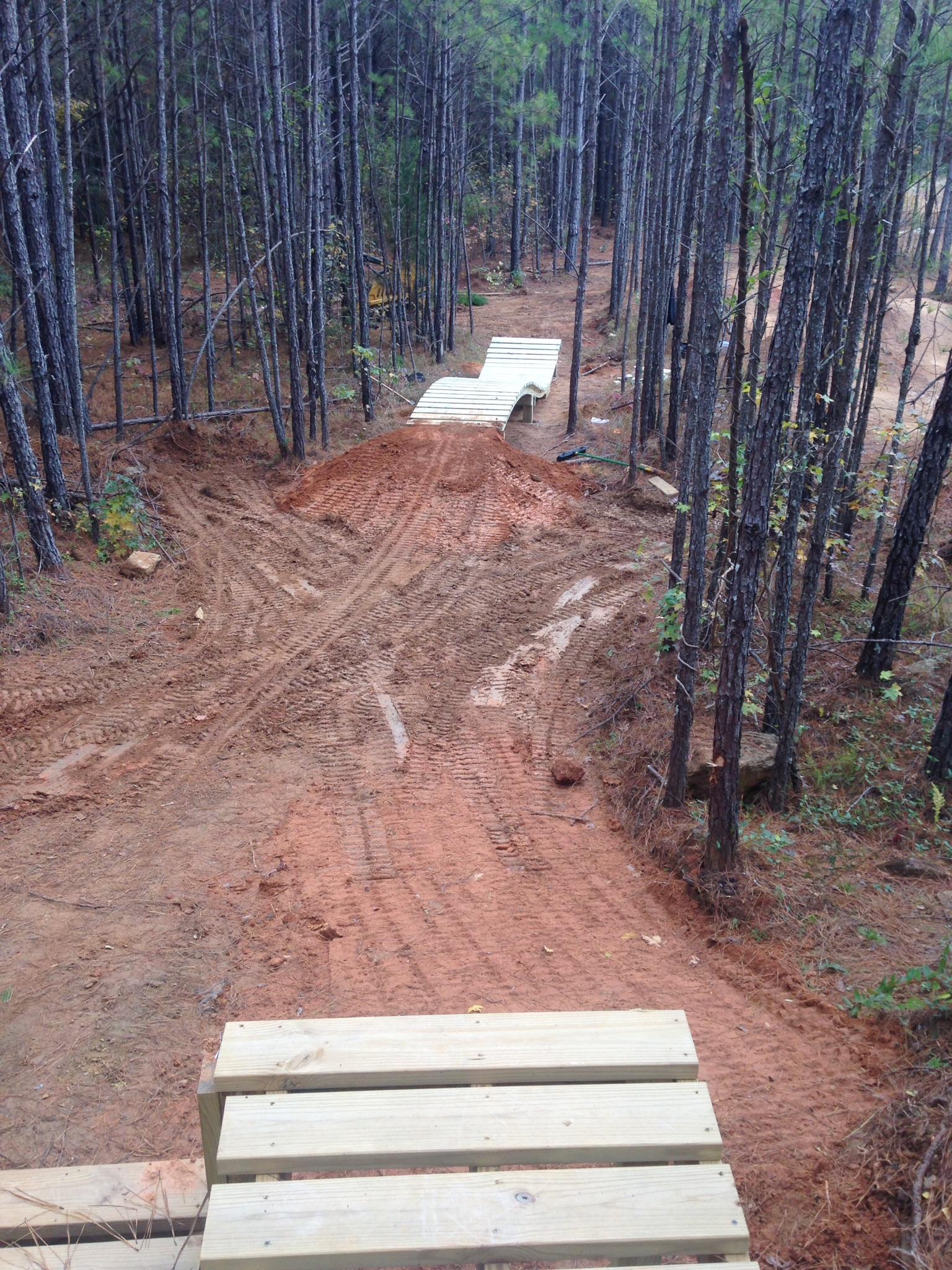 A dirt path through a wooded area featuring a wooden ramp or bridge in the foreground, leading over a dirt mound. The scene is surrounded by tall pine trees, with visible tire tracks and disturbed earth indicating recent activity. Allatoona Creek Park mountain bike trail.