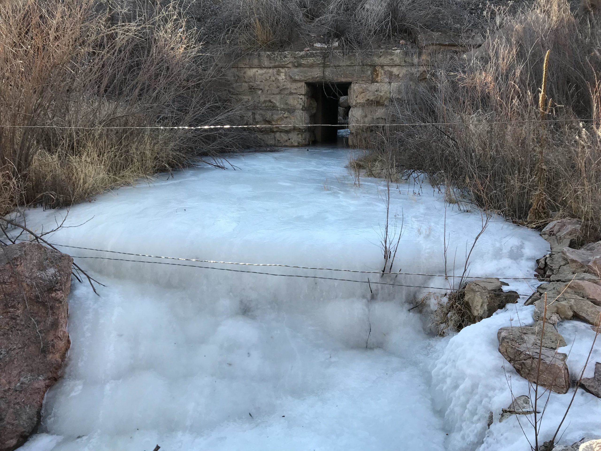 A frozen body of water flowing towards a stone structure, partially obscured by overgrown vegetation. The scene features icy textures and a few barren plants, with a wire fence running across the foreground. Santa Fe Trail mountain bike trail.