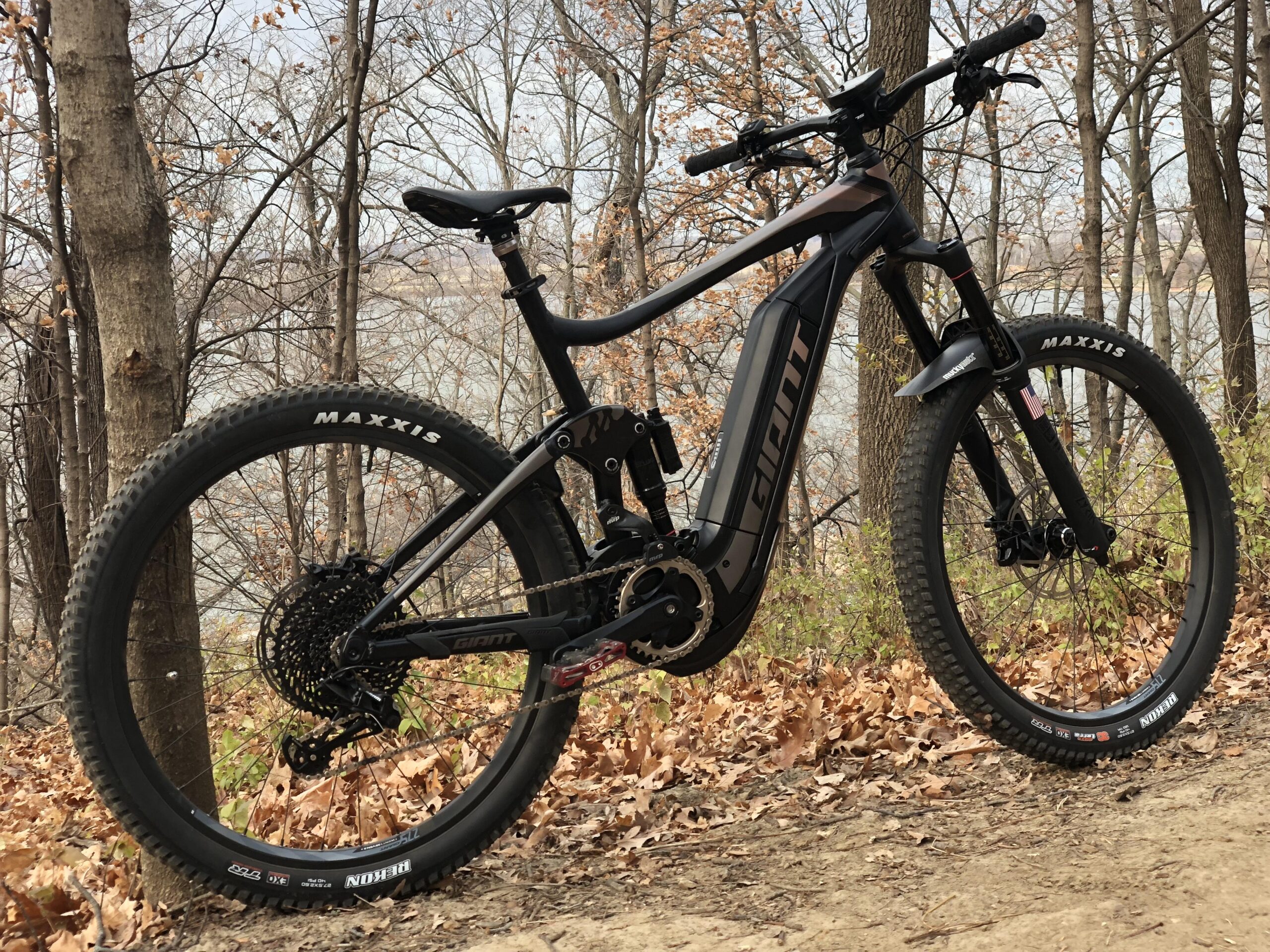 Giant FULL-E  0 SX PRO: A black electric mountain bike resting on a dirt path surrounded by bare trees and fallen leaves, with a river visible in the background. The bike features prominent Maxxis tires and a sleek design.
