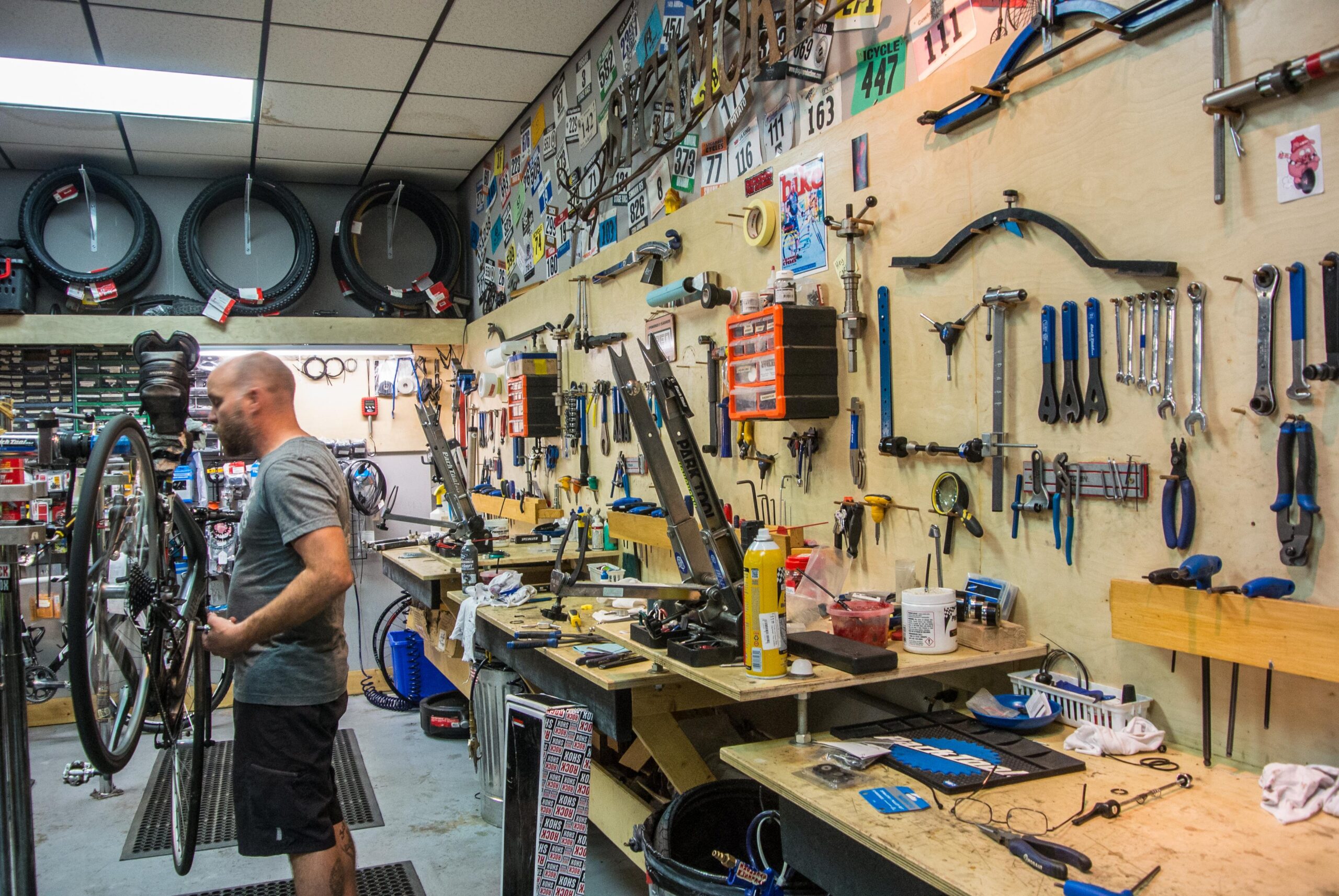 A bicycle mechanic holding a bike in a cluttered workshop filled with tools and equipment. The walls are adorned with various bike parts and license plates, and multiple workbenches are covered with tools, spare parts, and maintenance supplies.